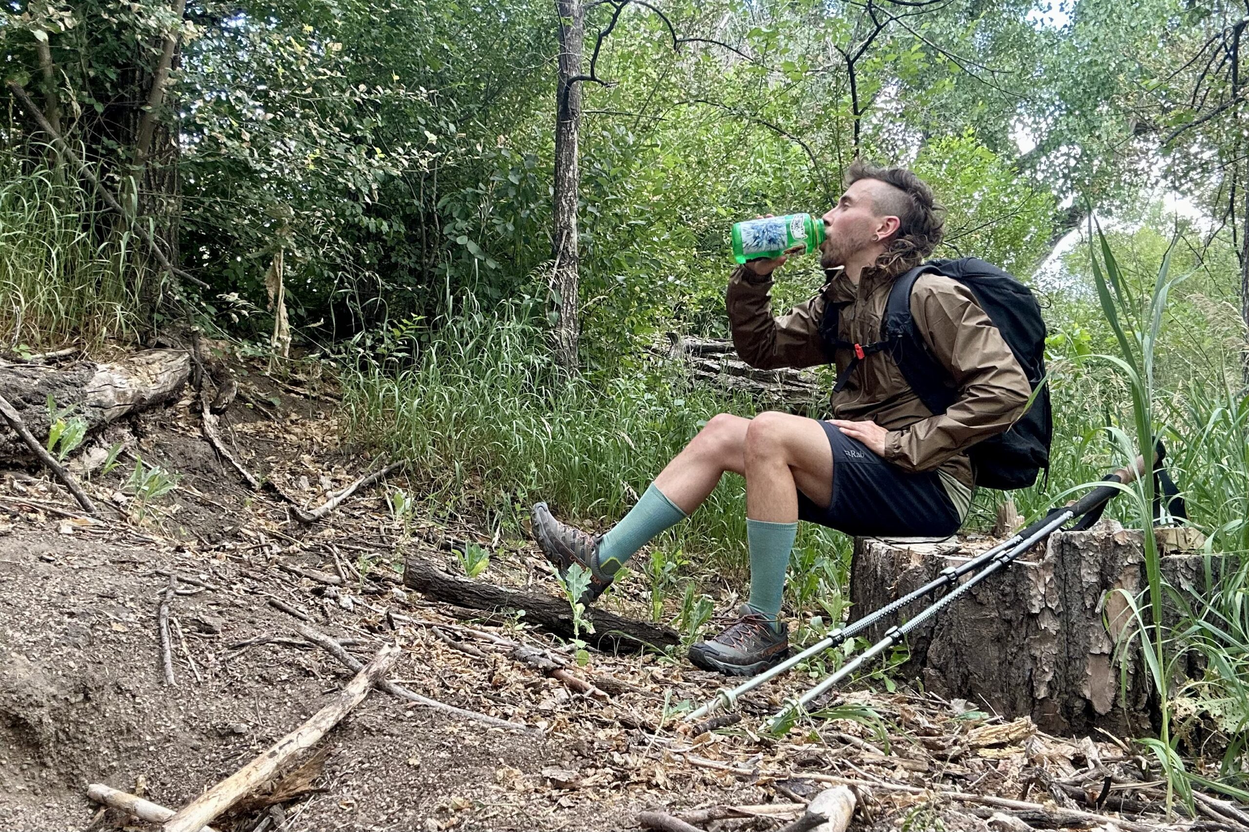 A hiker sits on a tree stump drinking water in a forest setting.