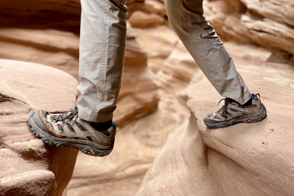 Man's legs from knee down steps across a deep gap between large sandstone boulders wearing the Merrell Moab 3 hiking shoes.