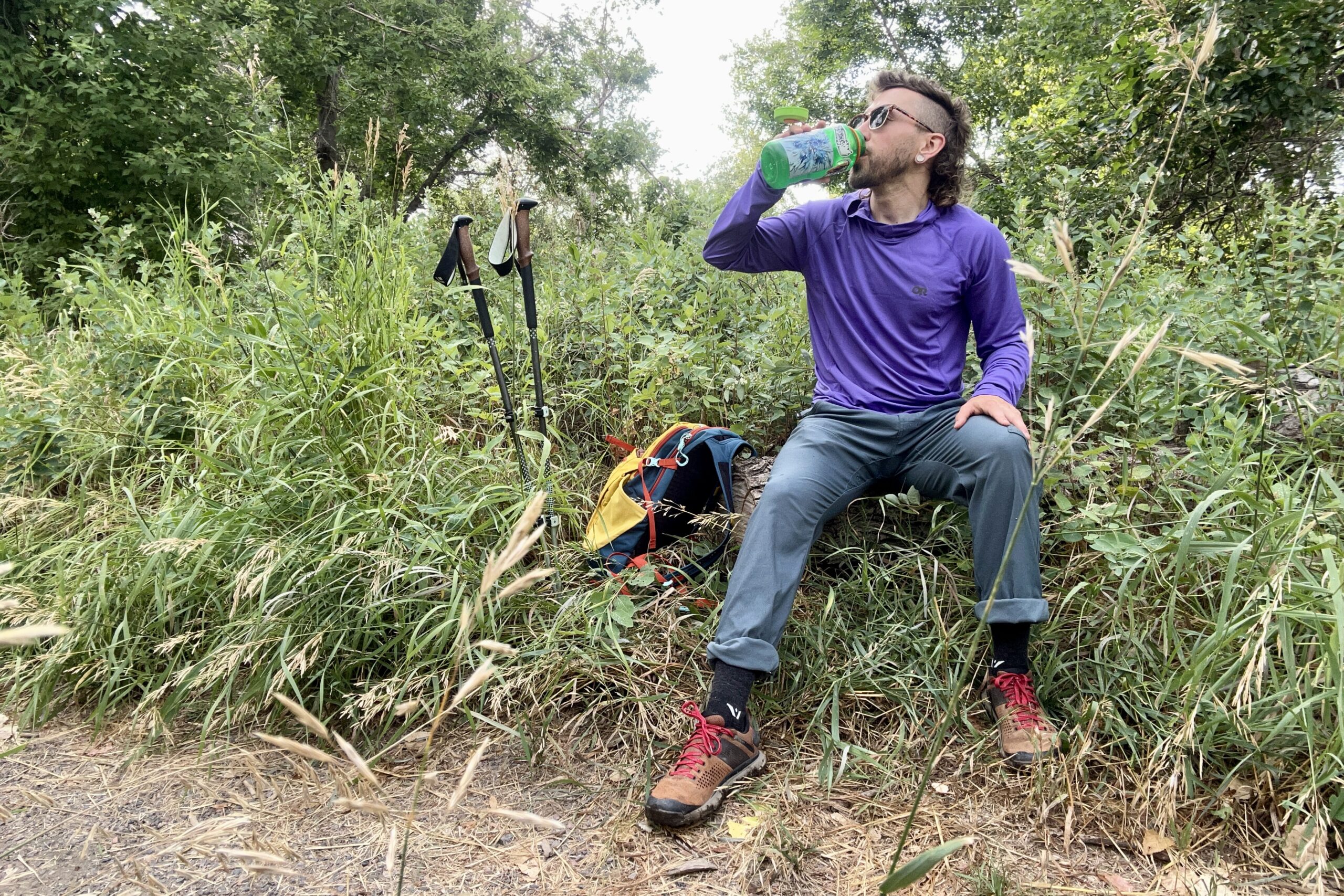 A man sits on a log drinking water on a hike with his pack and poles next to him.