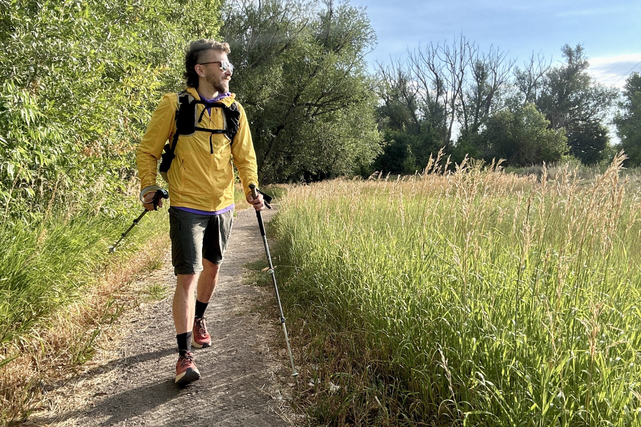 A male hiker looks off in the distance over a field of tall grass wearing a backpack and carrying poles.