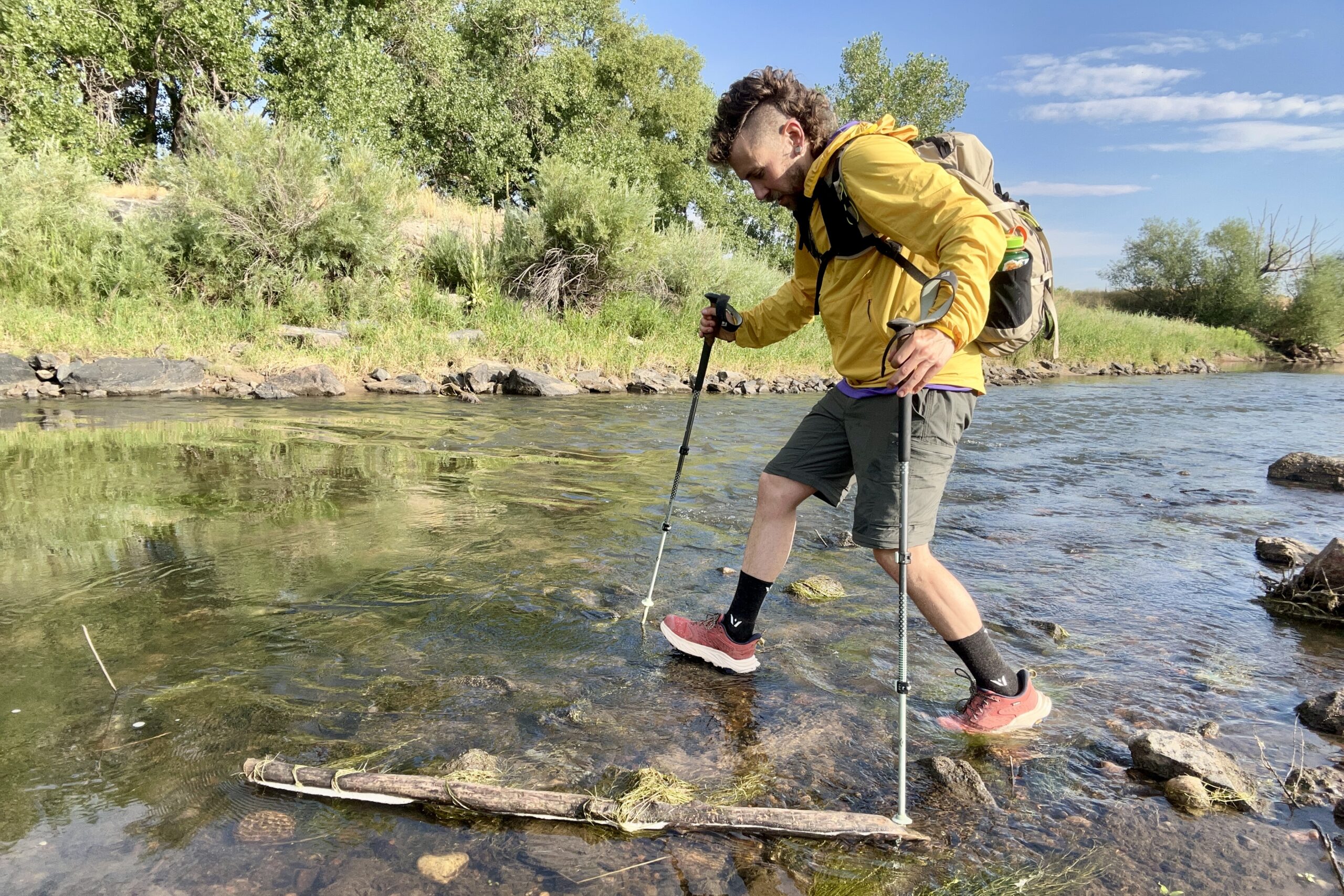 A male hiker walks across a river in a wilderness area using poles.