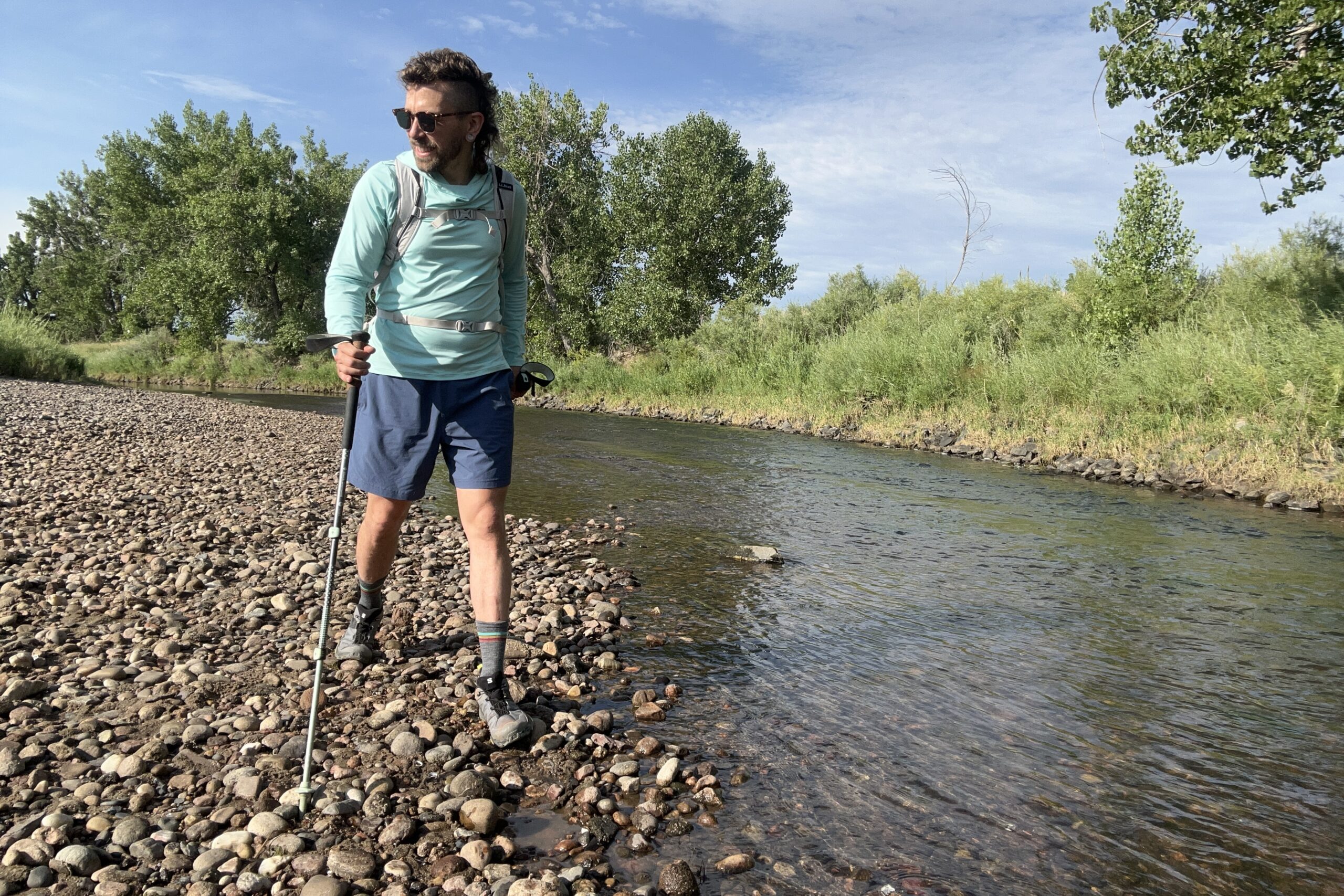 A hiker walks along a river on a bluebird day with poles and a backpack.