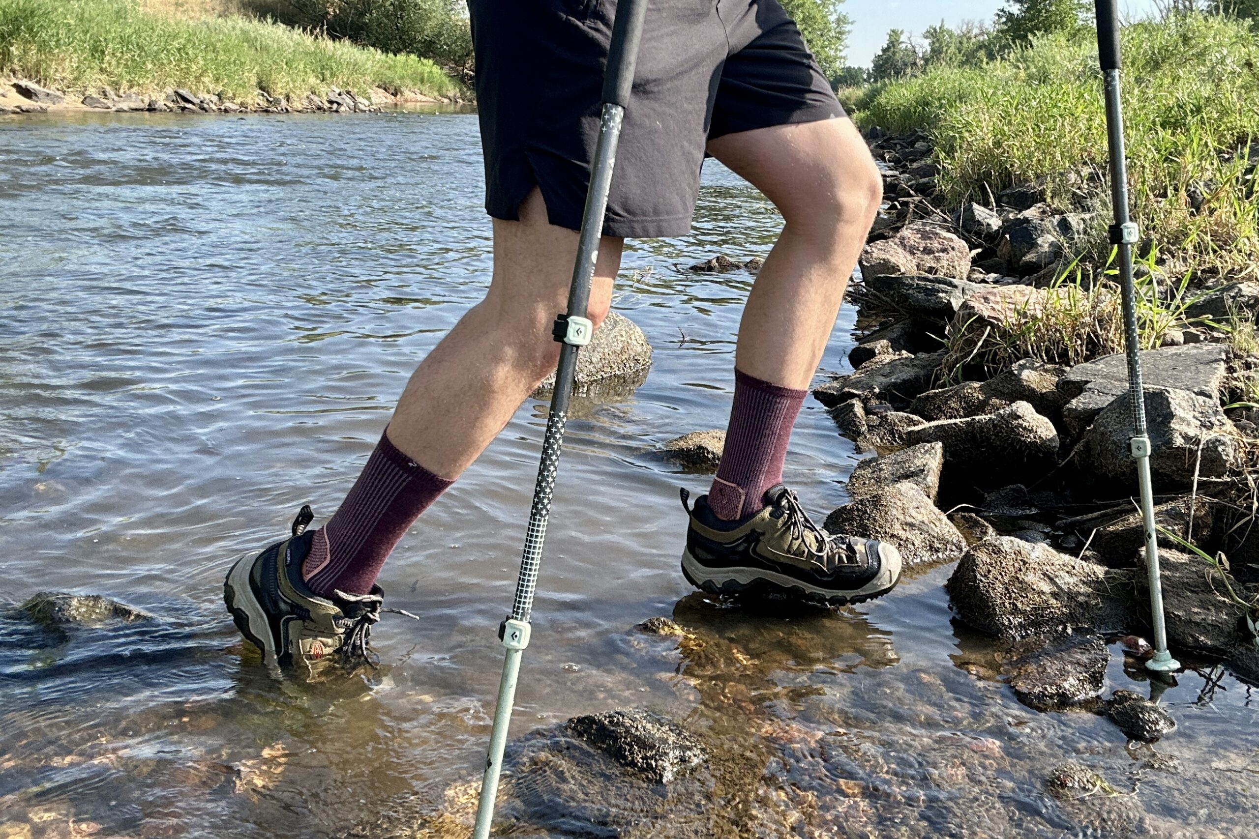 A male hiker crosses a river. The picture is from the waist down.