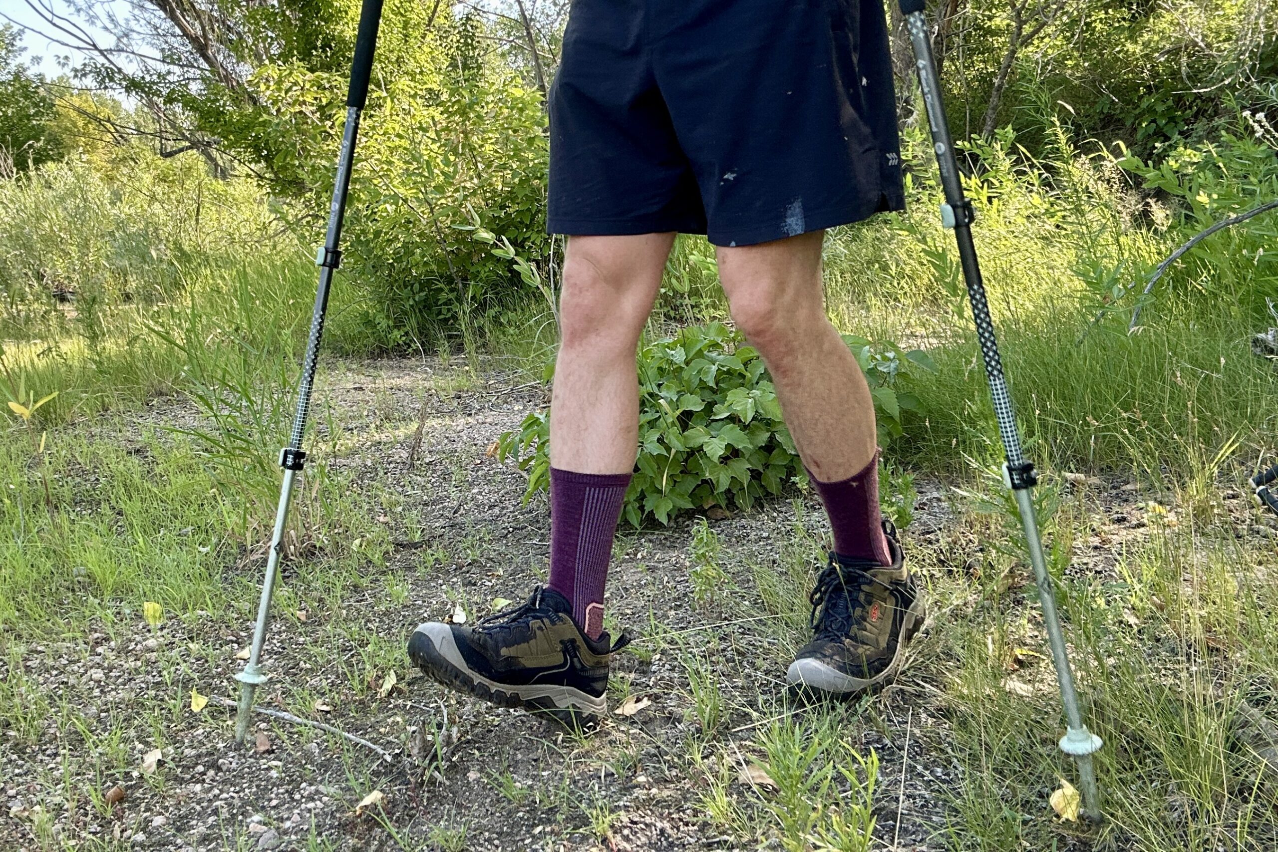 Uma foto de um homem da cintura para baixo caminhando por uma trilha usando meias na altura da panturrilha, shorts e calçados de caminhada.