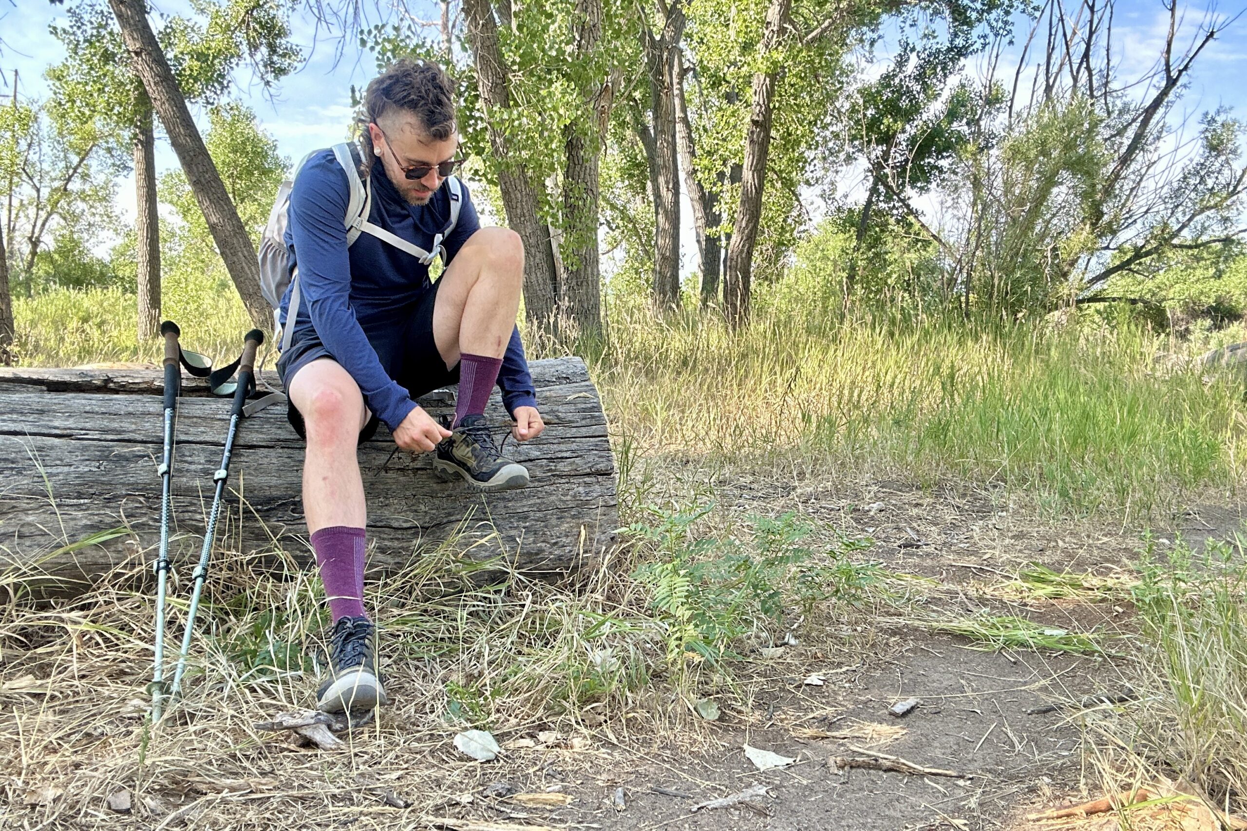 A male hiker sits on a log tying his shoes in a nature area wearing a blue sun hoody and black shorts under a blue sky.