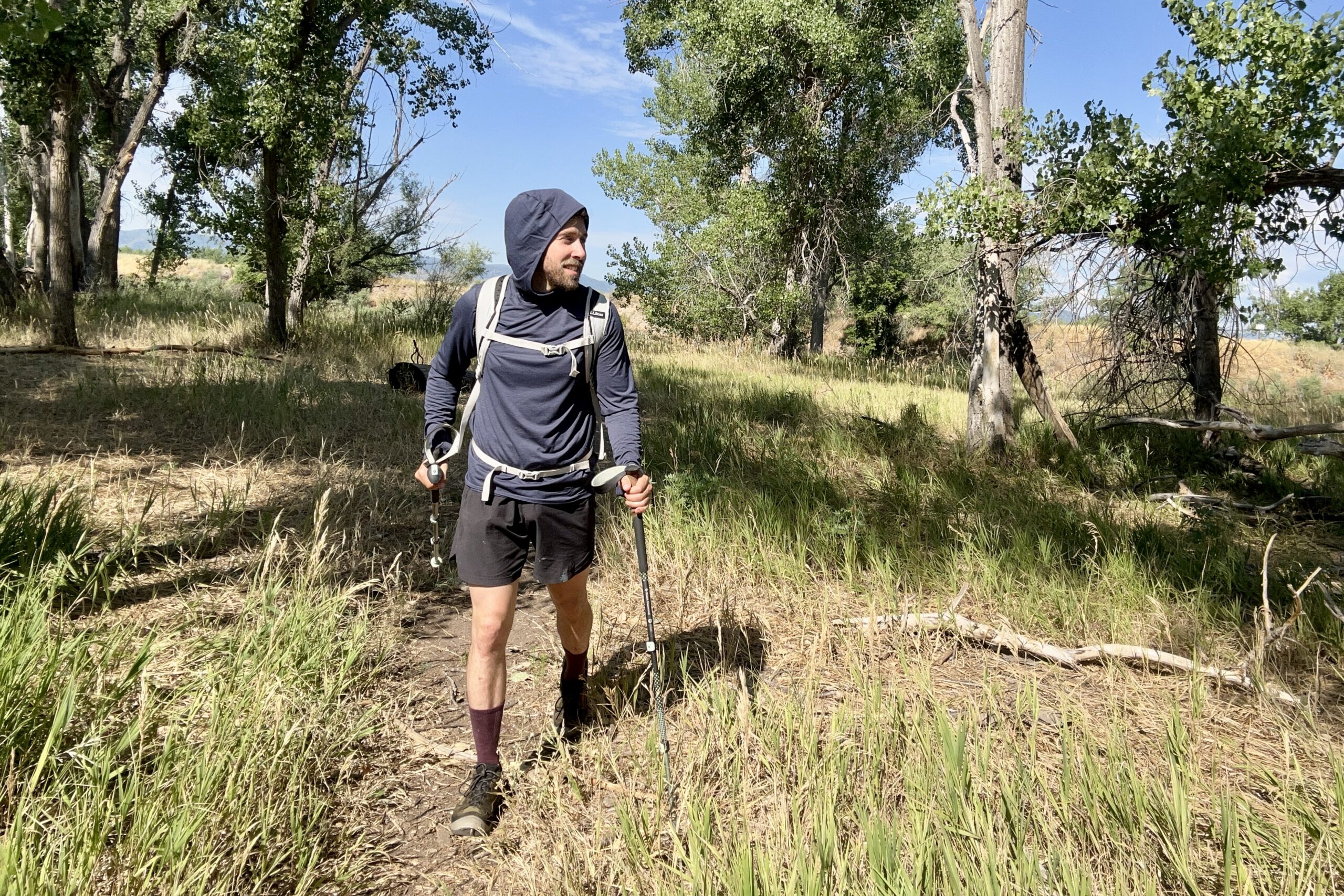 A male hiker walks through a nature area wearing a blue sun hoody and black shorts under a blue sky.