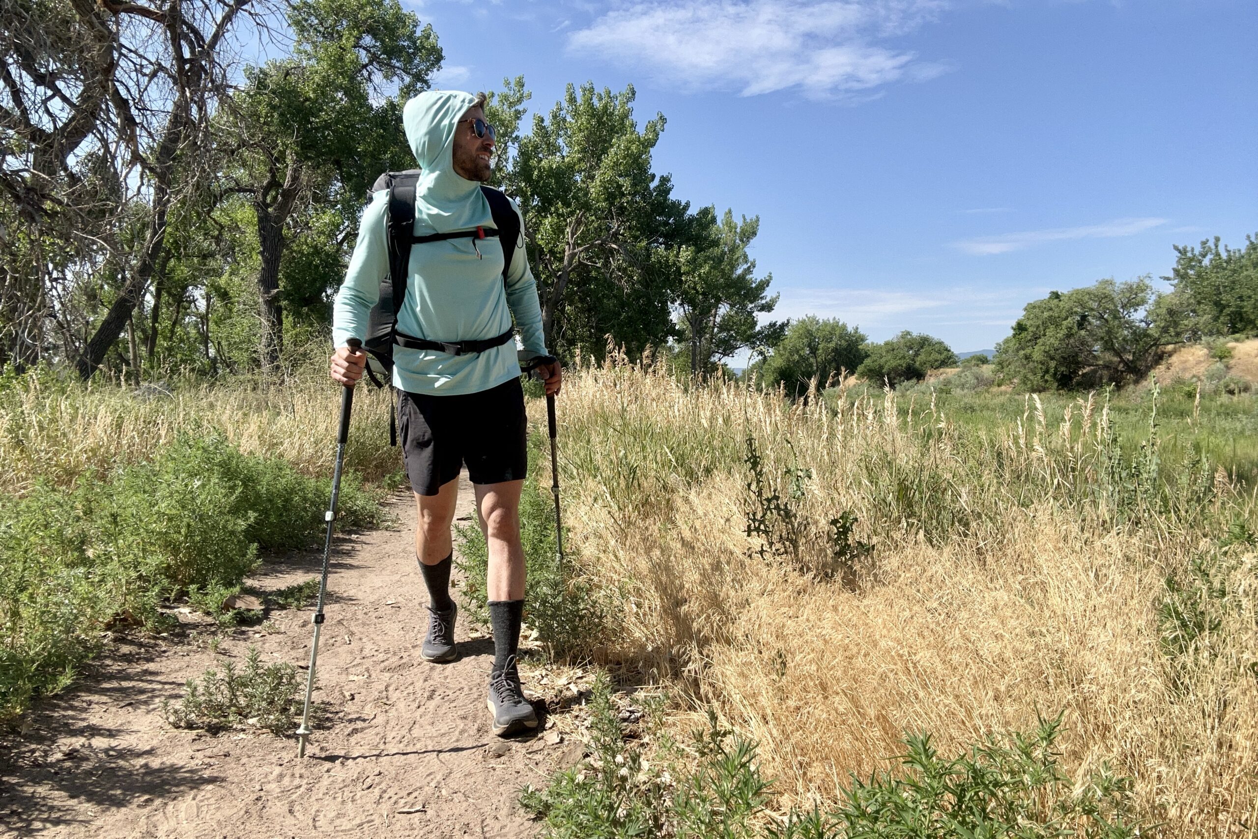 A hiker walks on a trail in a sun hoodie with a backpack and poles.