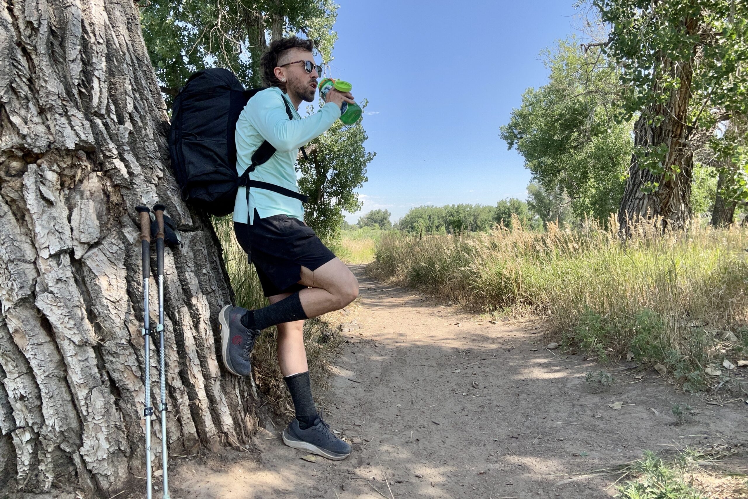 A hiker leans against a tree to stop and drink water on a trail in a sun hoodie under a blue sky.