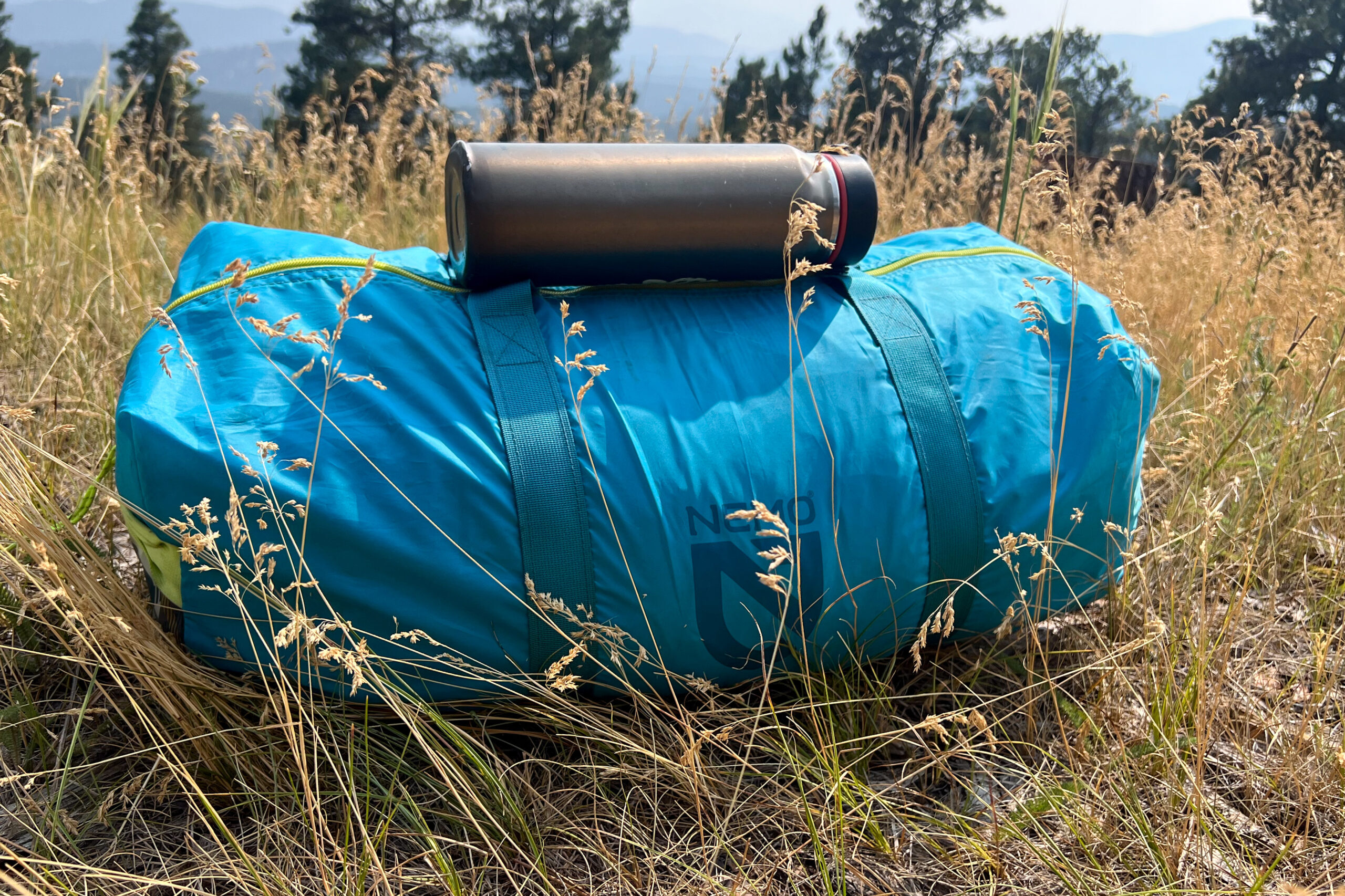 The image shows the Nemo Aurora Highrise tent inside it's stuff sack with an average 20 ounce water bottle sitting on top. The tent is in a grassy setting