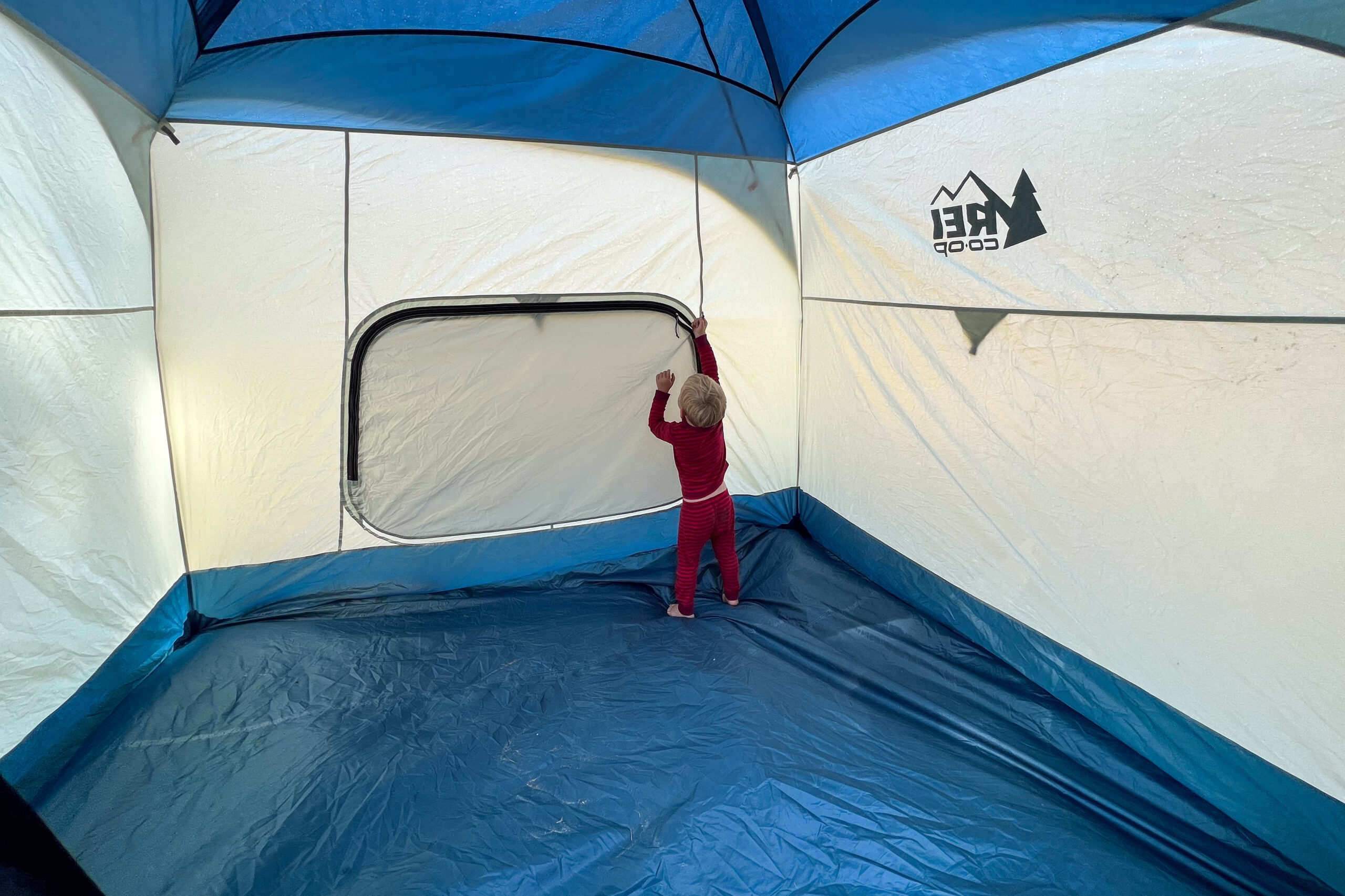 The image shows a child in an empty tent attempting to unzip the rear window of the tent.