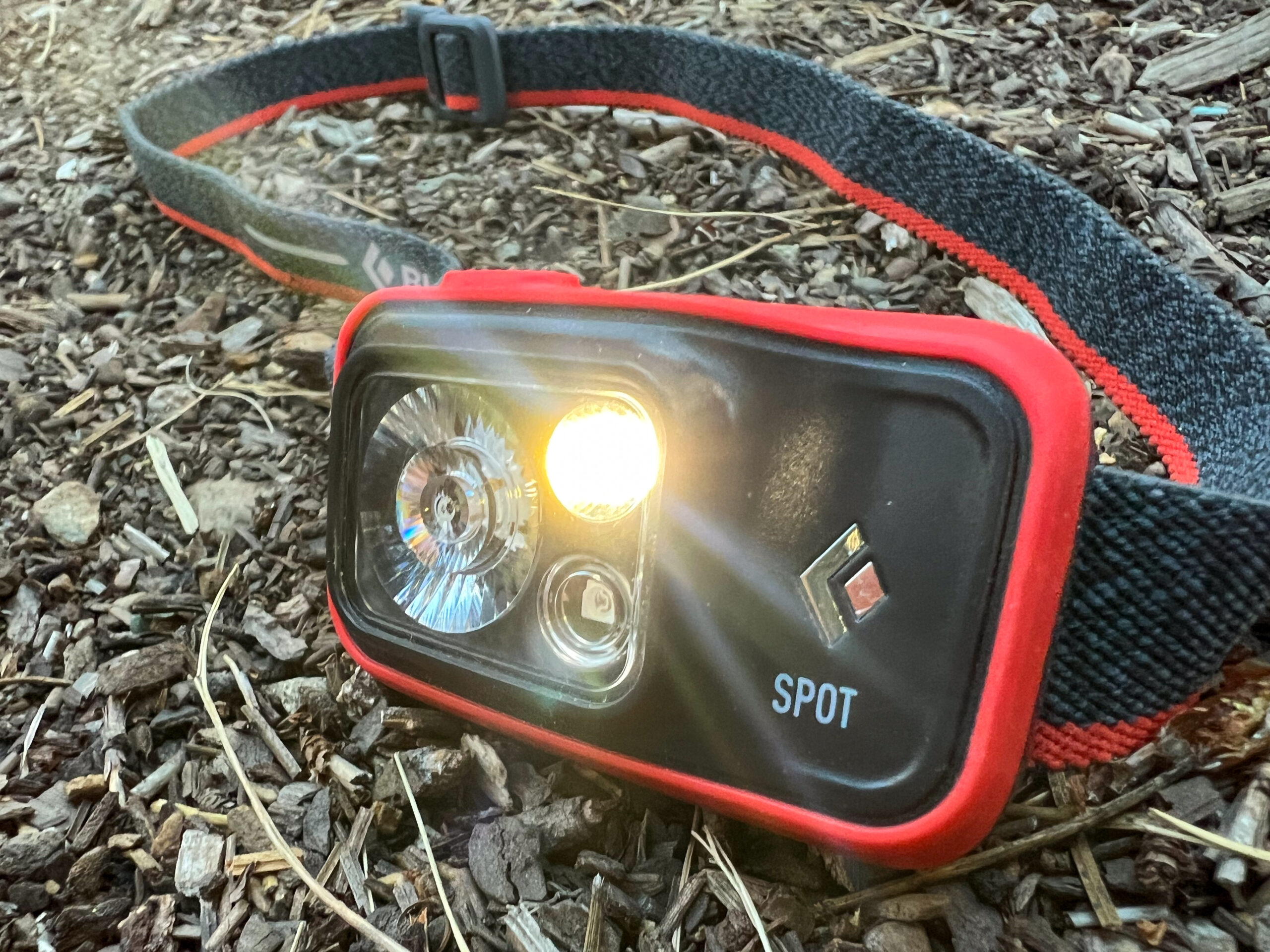 The image shows a red and black Black Diamond Spot 400 headlamp sitting on the forest floor. The light is on.