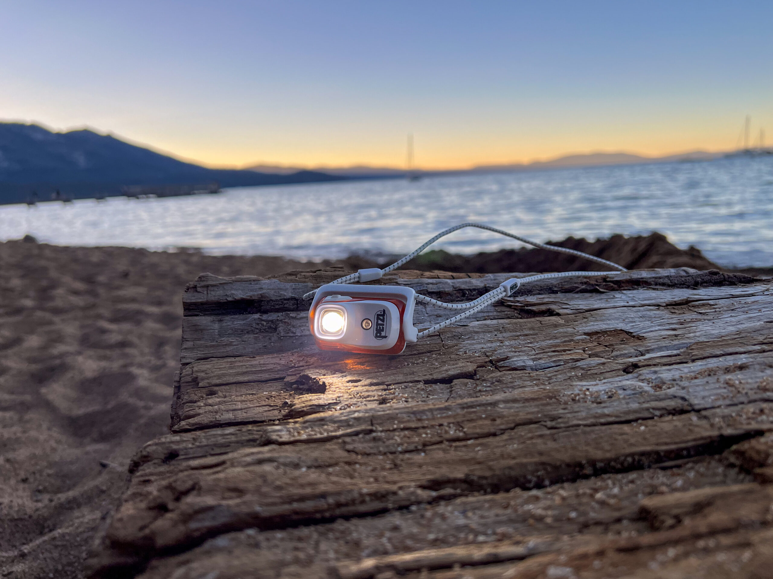 The image shows a headlamp sitting on a piece of driftwood and a beach with the sun setting in the background.