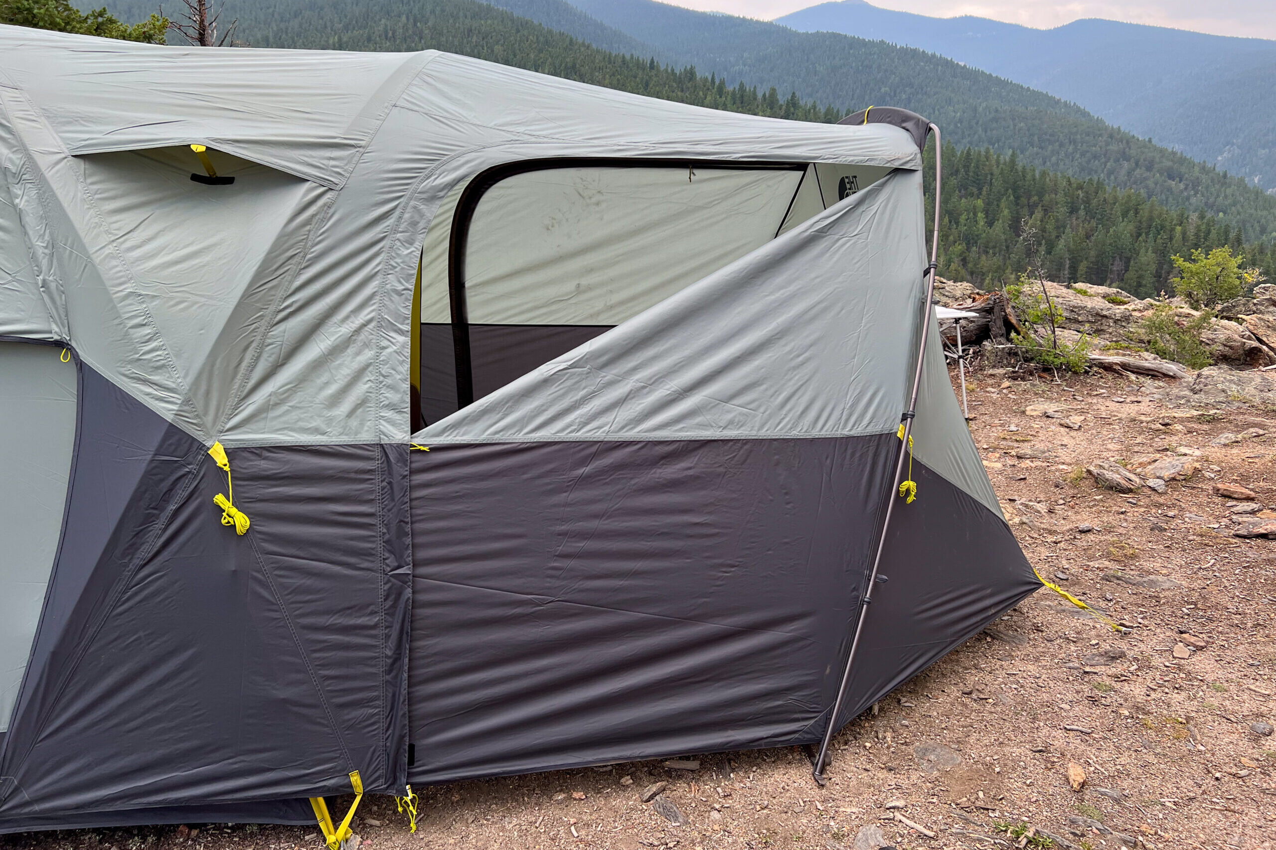 The image shows The North Face Wawona 6 tent from the side. The from door of the vestibule is cracked and the kickstand style vent on the rainfly is open to allow airflow into the tent. There are mountain in the background and the sky is showing signs of rain