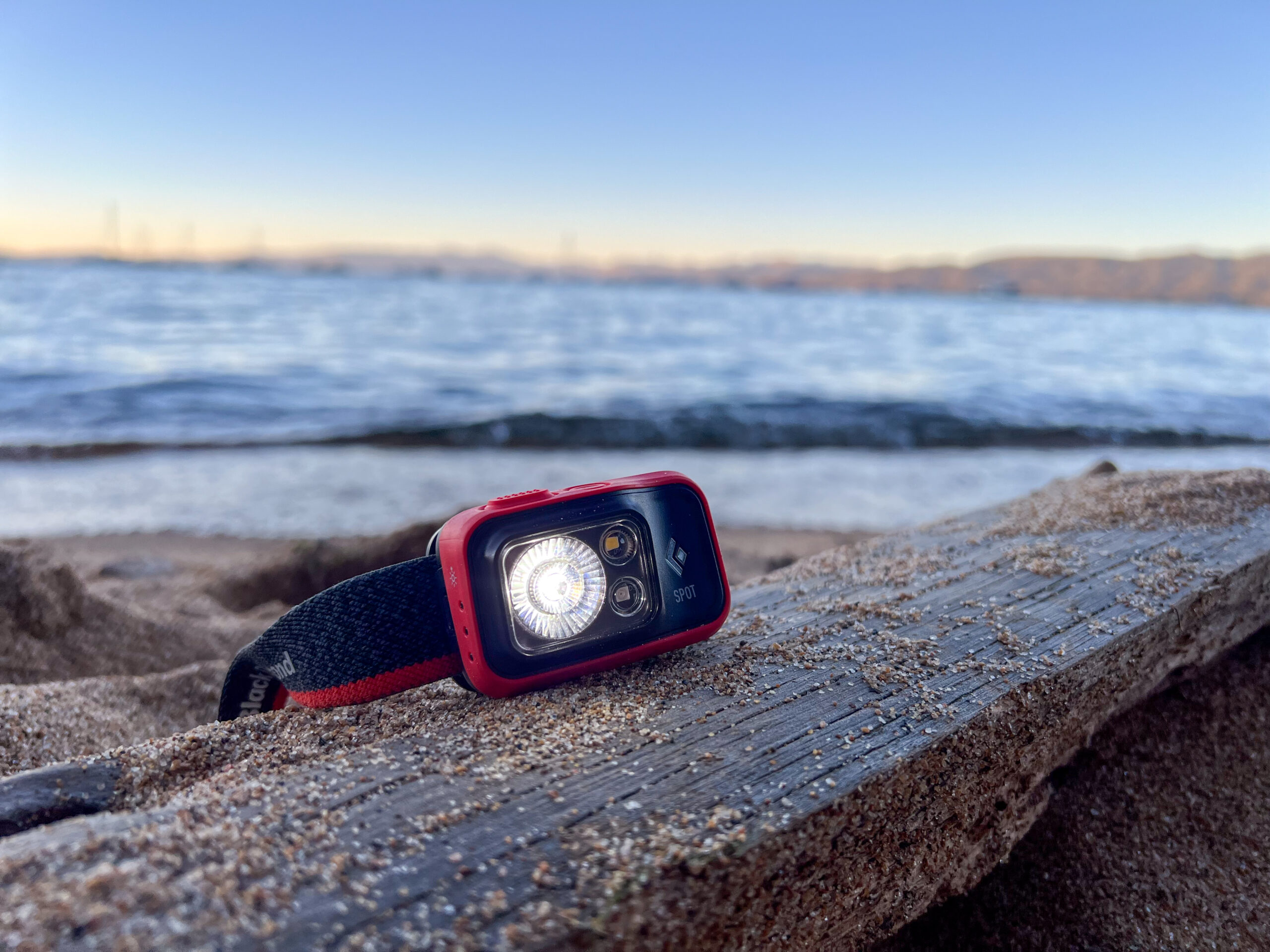 The image shows the Black Diamond Spot 400 sitting on a beach with it's floodlight illuminated. There is water in the background.