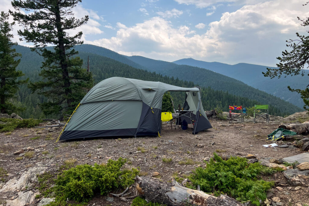 The image shows a wide shot of The North Face Wawona 6 tent. Both front vestibule doors are open and rolled up to allow a full view through the open vestibule. There are tree covered hills in the background. The sky is blue with mixed in clouds.