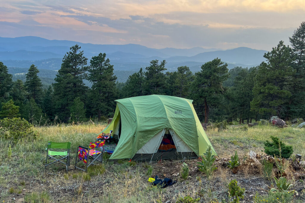 A medium wide photograph of the REI Wonderland 4 camping tent. The ten is set on a mountainside and the sun is setting in the background. Wildfire smoke shrouds the sky and the sun is setting with a twinge of orange in the clouds.