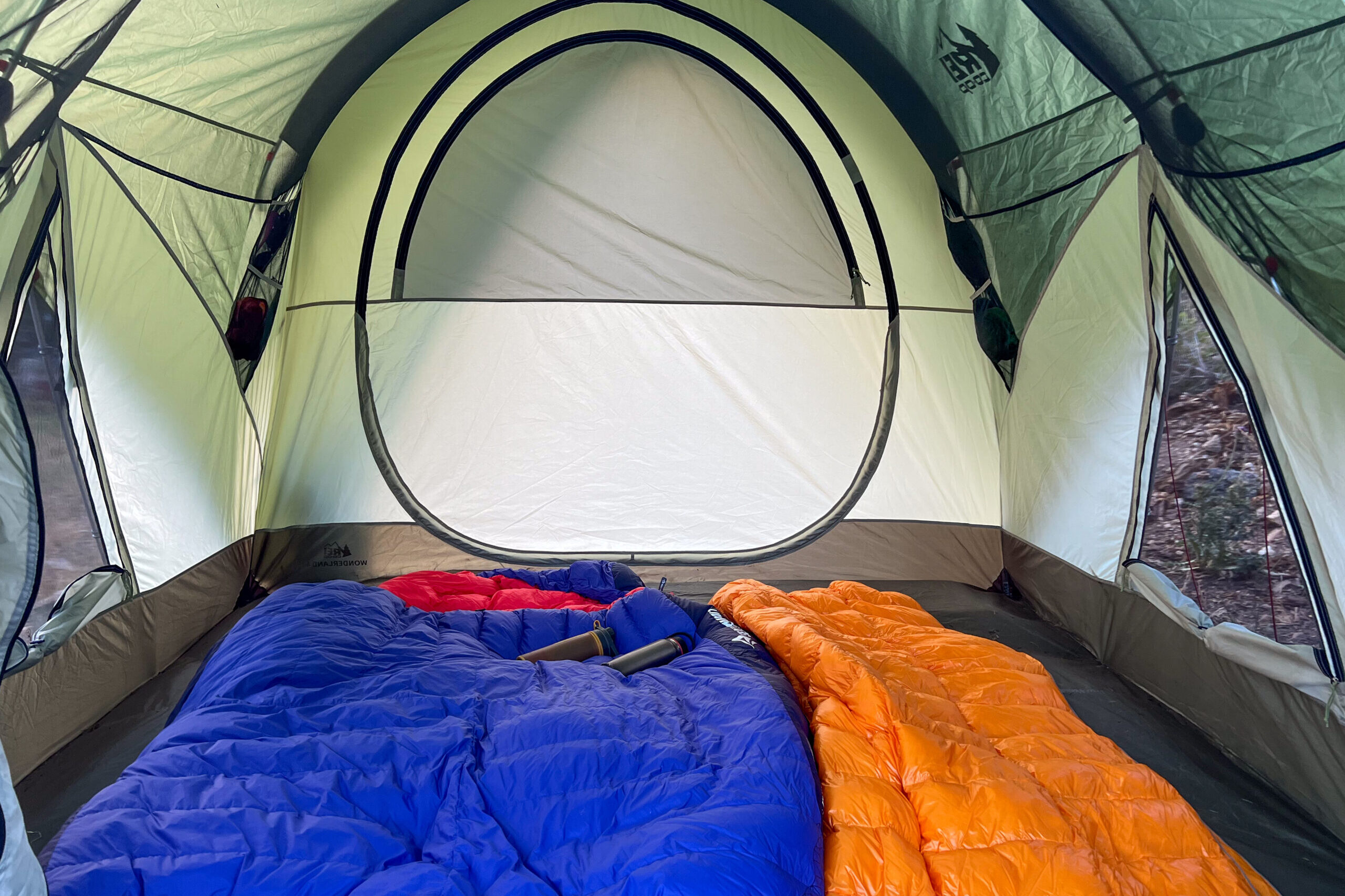 The image shows the inside of the REI Wonderland 4 tent, There is a double sized sleeping bag on the left and a single sized sleeping bag on the right. There isn't much room for another sleeping bag. Both windows one either side of the tent are rolled down and stowed.