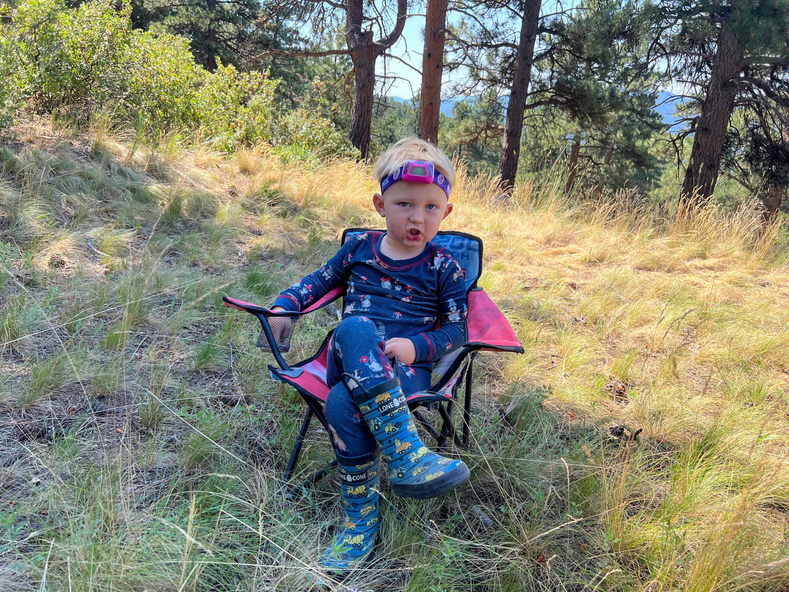 The image shows a child wearing a headlamp. They are sitting in a small camp chair with their legs crossed.