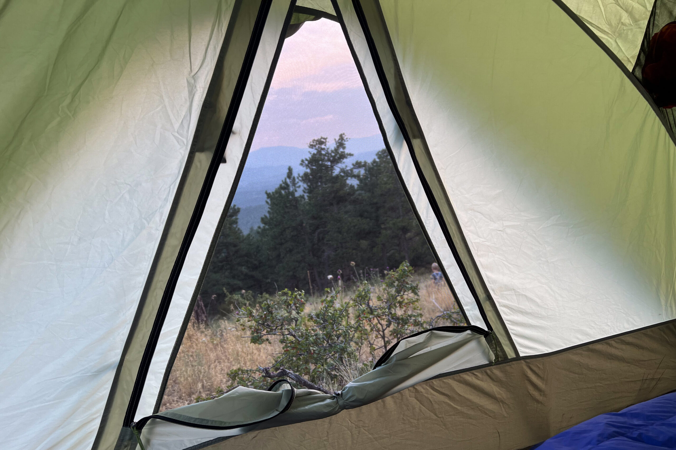 Image from inside the REI Wonderland 4 tent. The image shows one of the internal windows rolled down and stowed. The view is predominantly through the mesh window. The sun is setting, there are mountains in the background, and the sky is purple and orange.