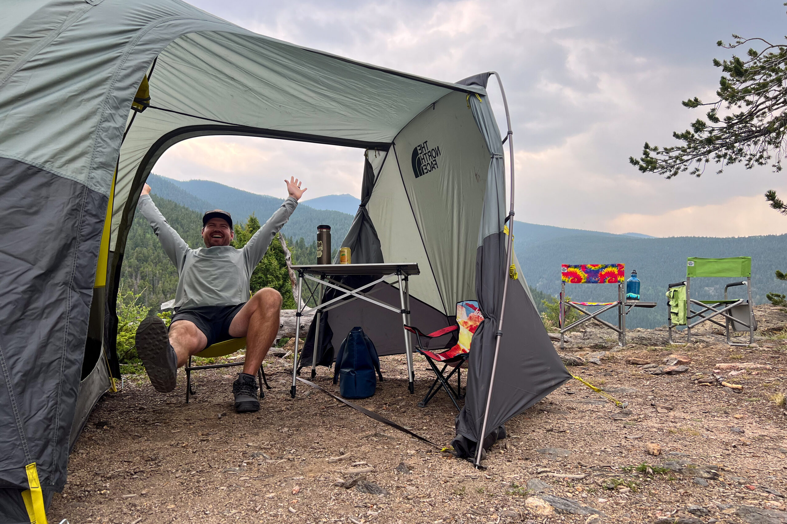 The image shows a camper sitting in a chair inside the front vestibule of The North Face Wawona 6 tents. The camper is smiling and his hard are in the air. The sky is ominous with rain clouds and there are mountains in the distance.