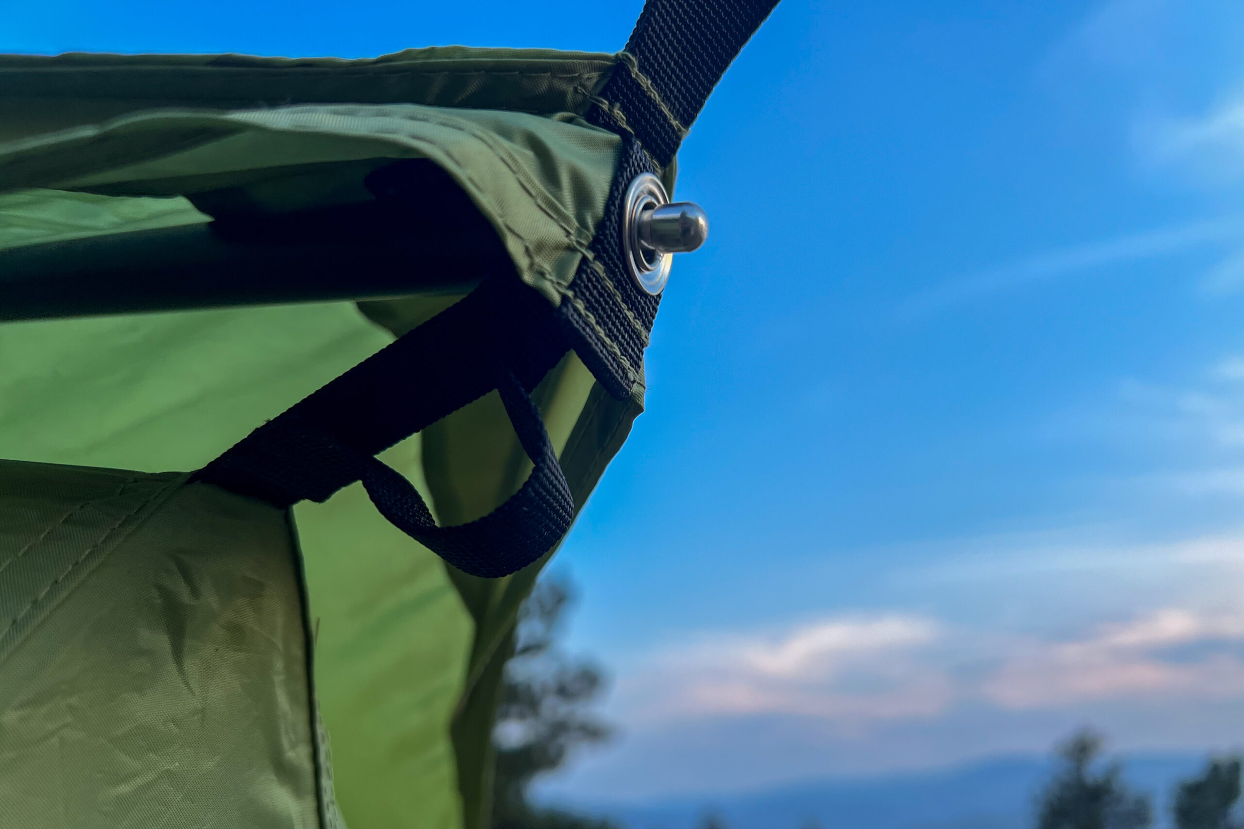 A picture of how the REI Wonderland tent rainfly attached to the top pole. The image shows the top pole fitting into a metal grommet. The sun is setting and the sky is deep blue with wispy clouds in the background.