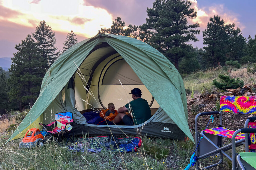 A father and son sit inside the REI Wonderland 4 tent. The sun is setting and the sky is vibrant orange and purple.