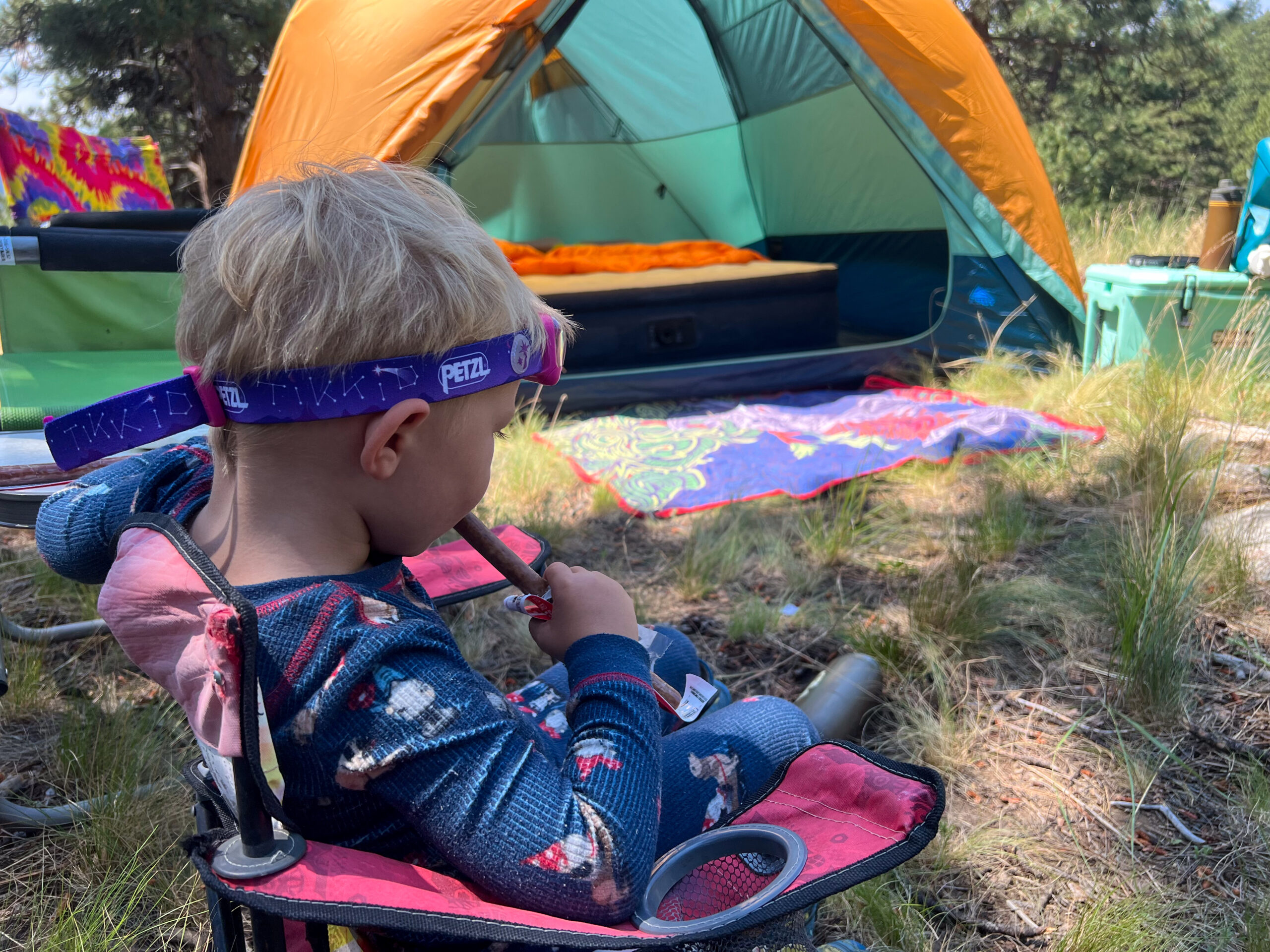 The image shows a child having a snack and wearing a headlamp. The child is facing away from the camera and sitting in front of a tent.