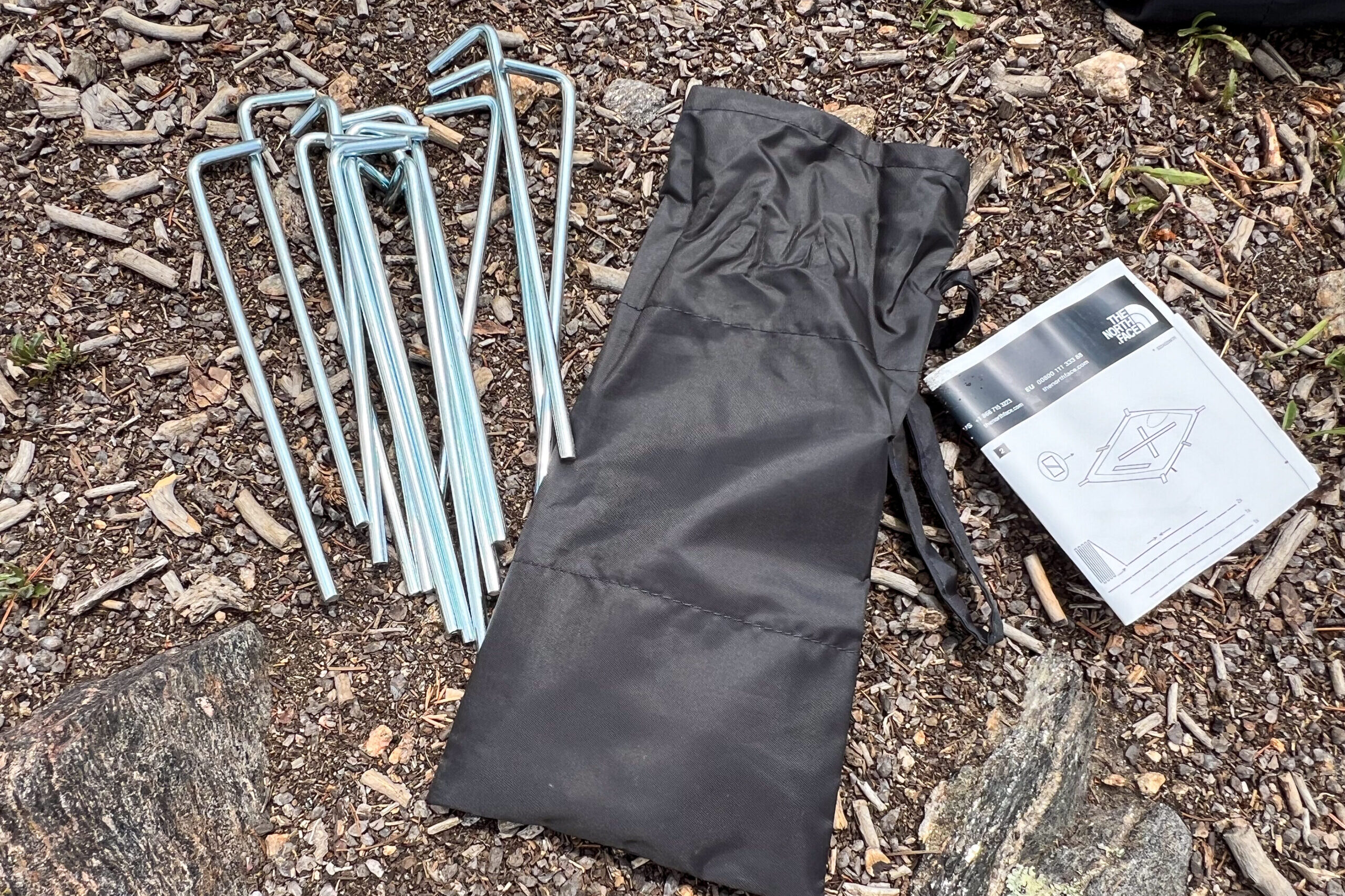 The image shows 12 tent stakes, a stake bad, and a set of instructions for The North face Wawona 6 tent. The collection of items is laying on the forest floow.