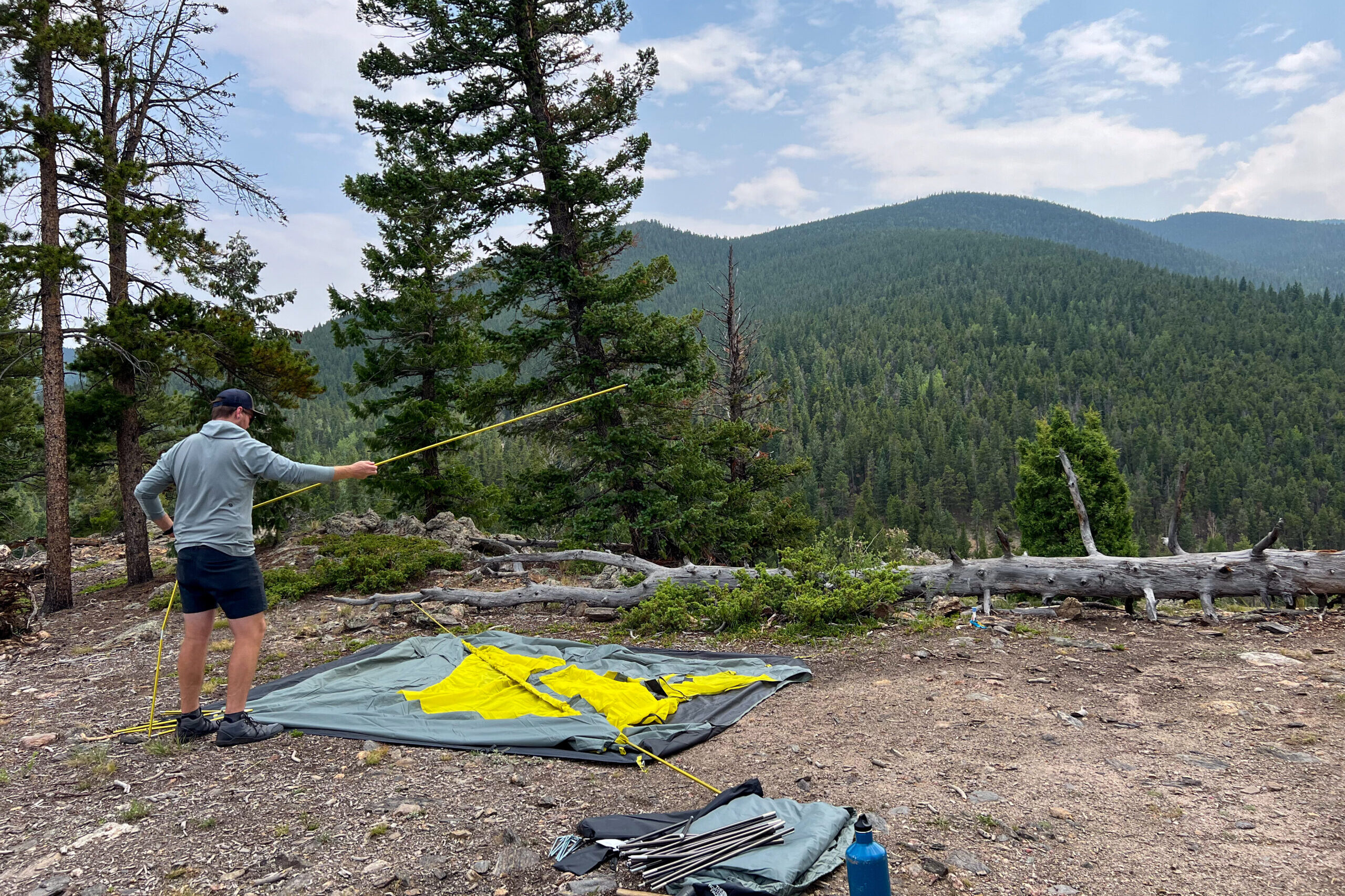 The image shows a male camper setting up The North Face Wawona 6 tent in a mountainous setting. The camper is connecting a yellow tent pole and the tent body is laying flat on the ground.