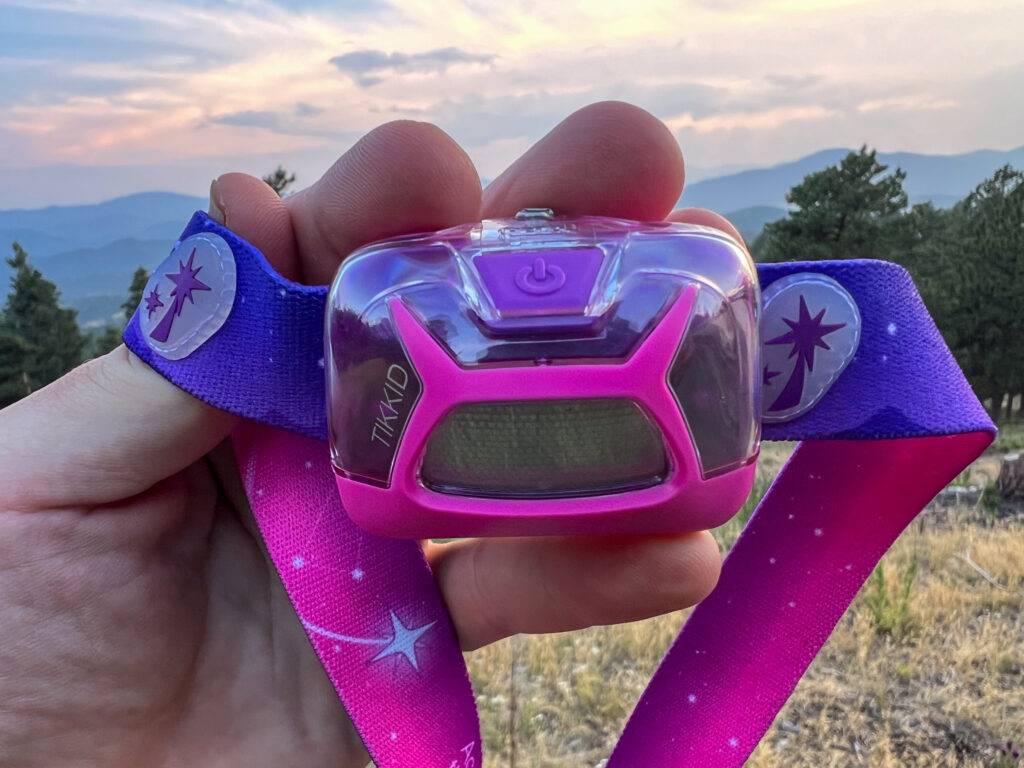 The image shows a headlamp in the hand of a camper. The headlamp is facing the camper and there are mountains in the background.