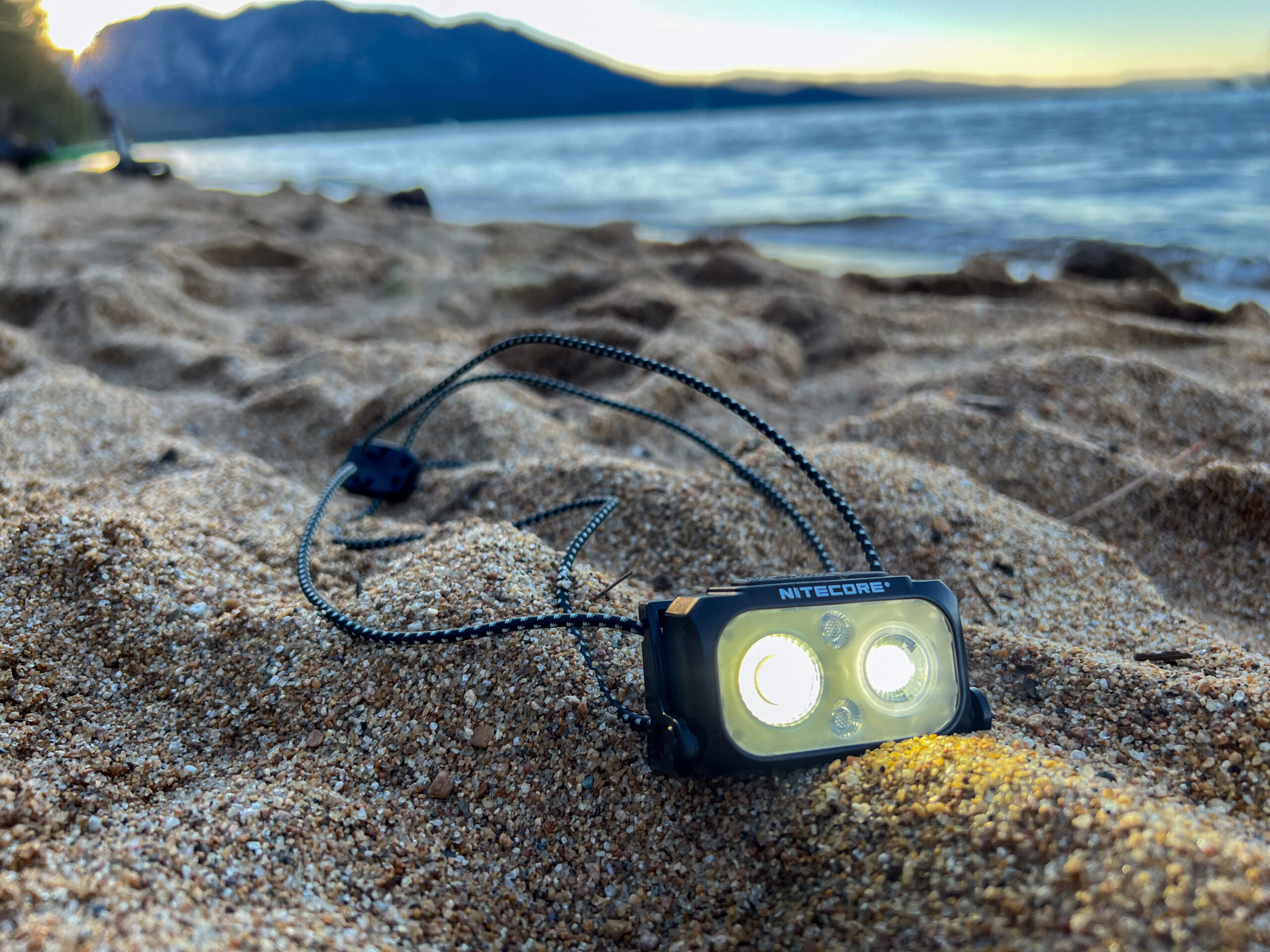 the nitecore headlamp aglow resting on the sand next to a large body of water with snow dusted mountains and sun setting behind it. 