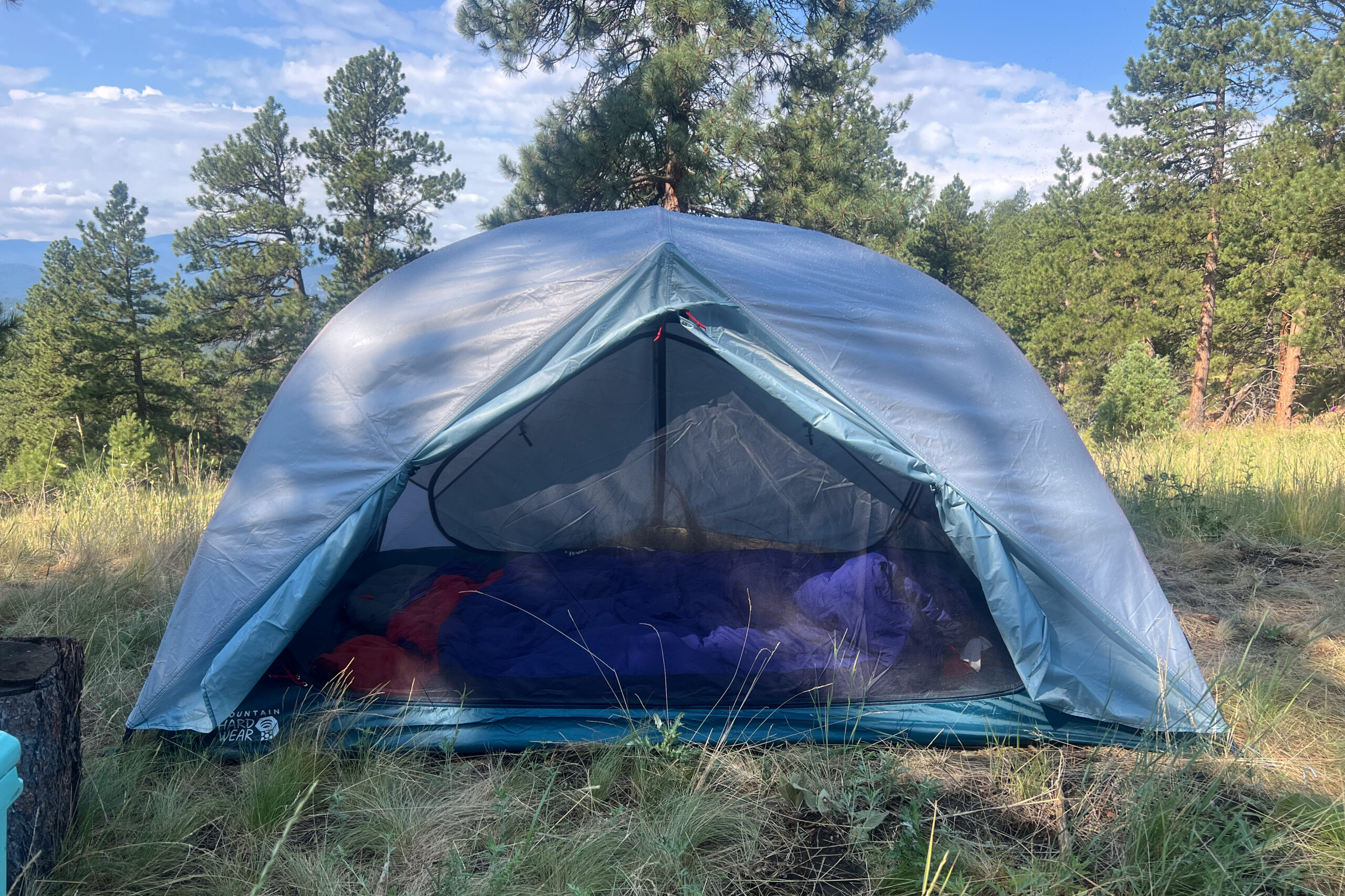 The Image shows the Mountain Hardware Mineral King 3 from the front with the front vestibule rolled back. The tent is in a grassy setting and surrounded by trees.