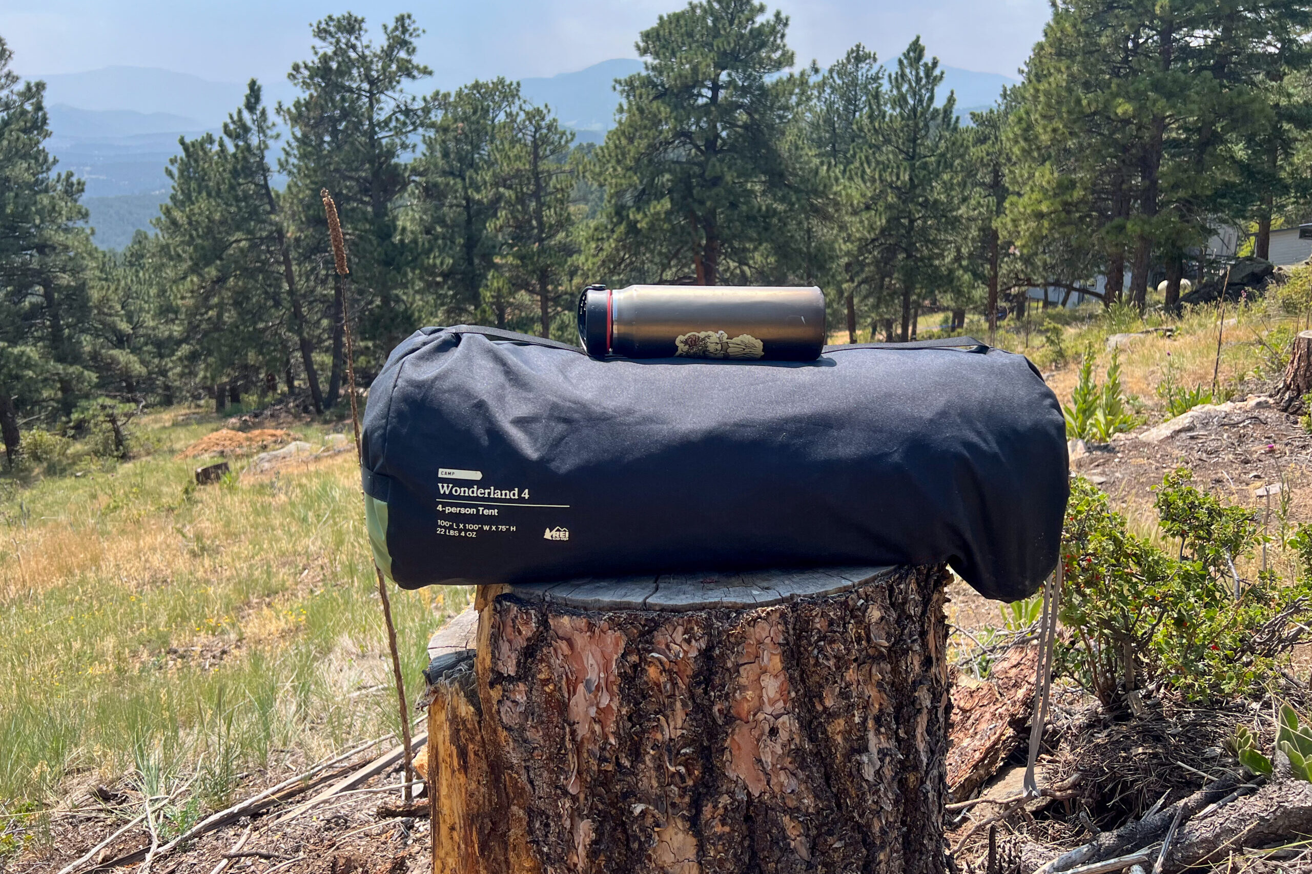 A photo of a standard size water bottle on top of the the REI Wonderland 4 tent in it's original stuff sack to show how large the tent is. The ten is sitting on top of a tree stump with ponderosa pine trees in the background with larger mountains in the distance.