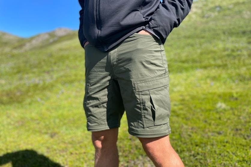 A man stands in the alpine with hands in pockets while wearing cargo shorts