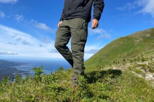 A man walking in the alpine wearing green pants with blue sky behind.