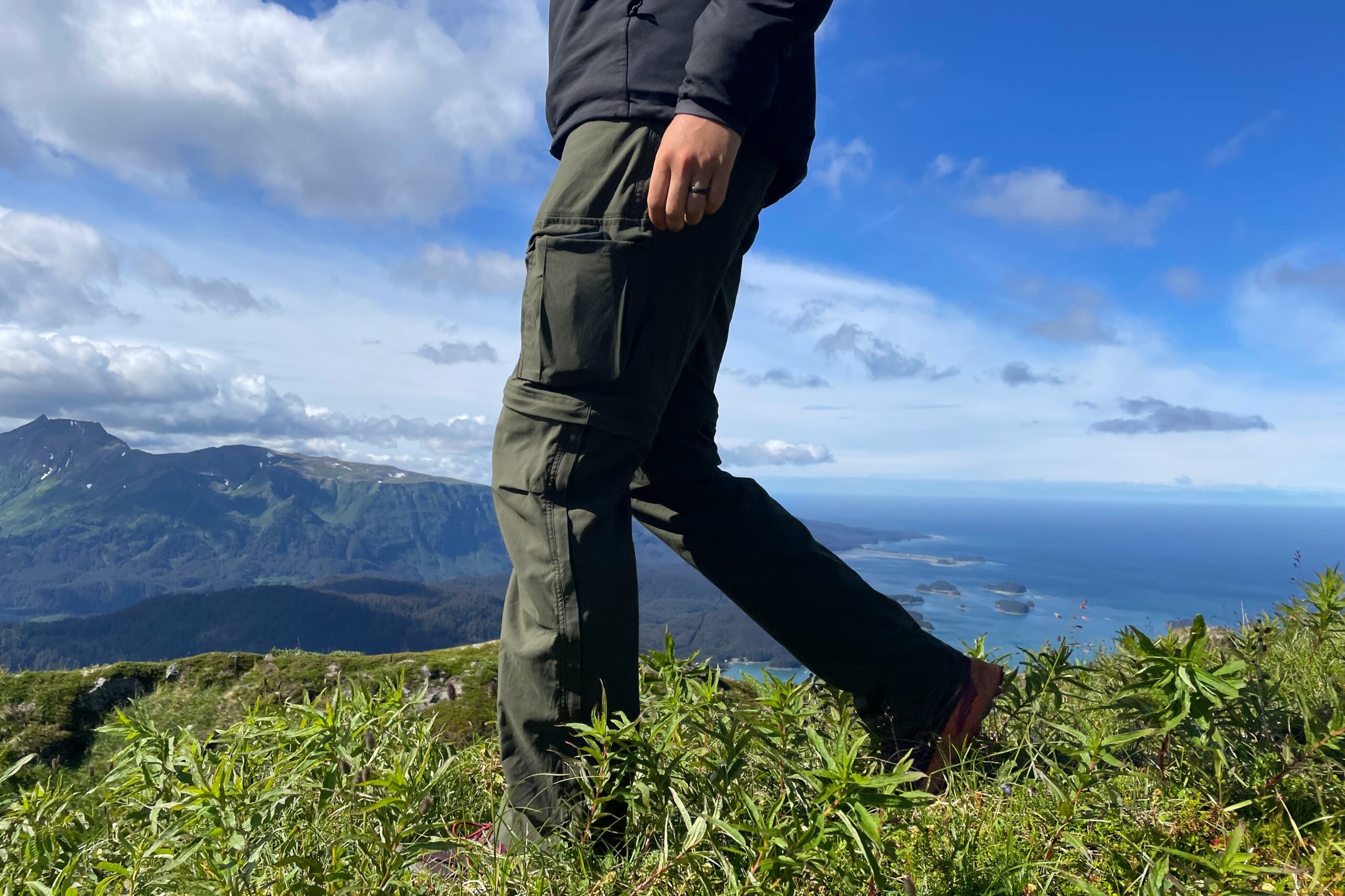 A man walking to the left through green grass with a view of the ocean in the background.