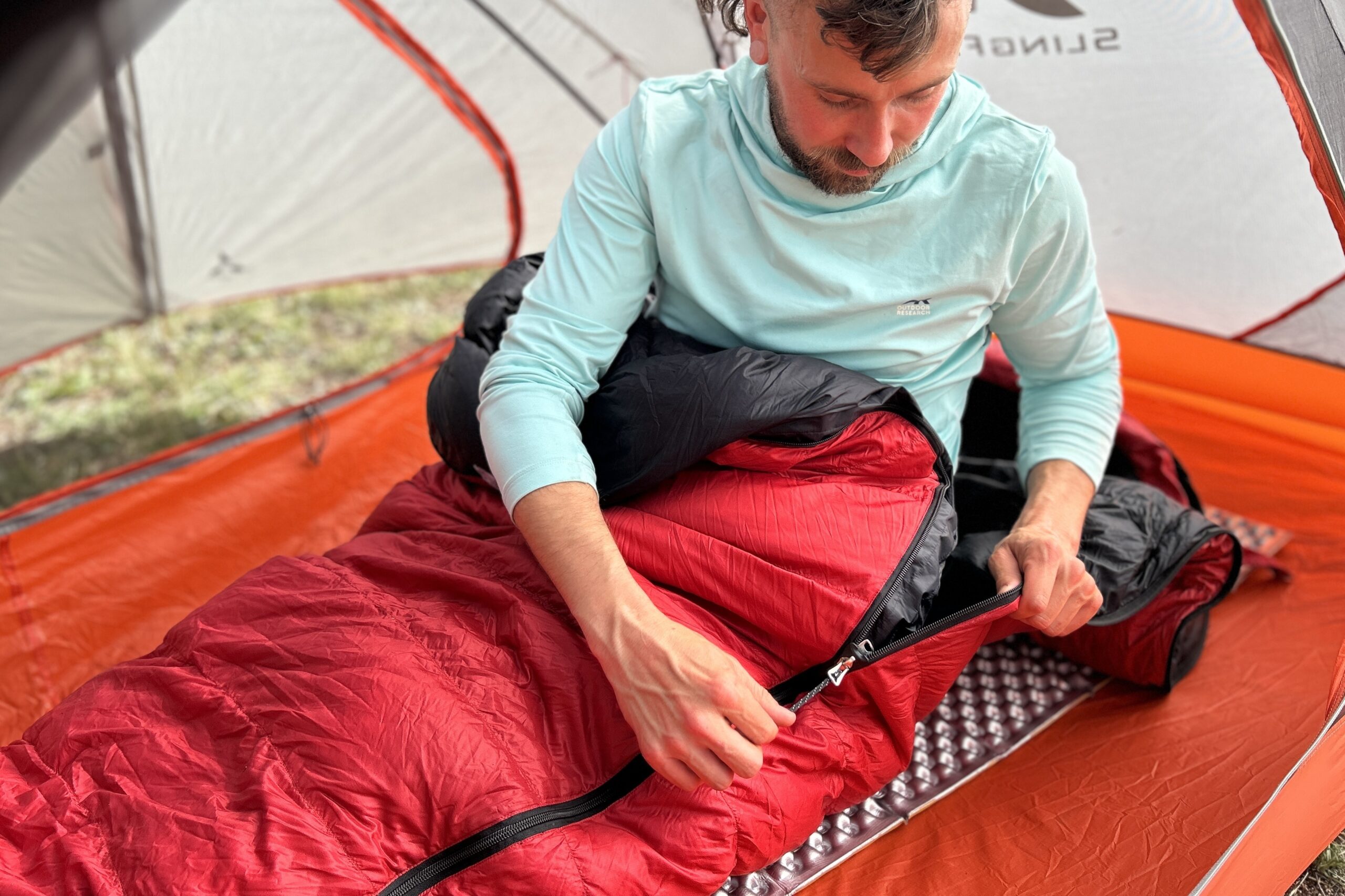 A man sits up on a pad and is in a sleeping bag from the waist down. He messes with the zipper on a red sleeping bag inside of a tent.