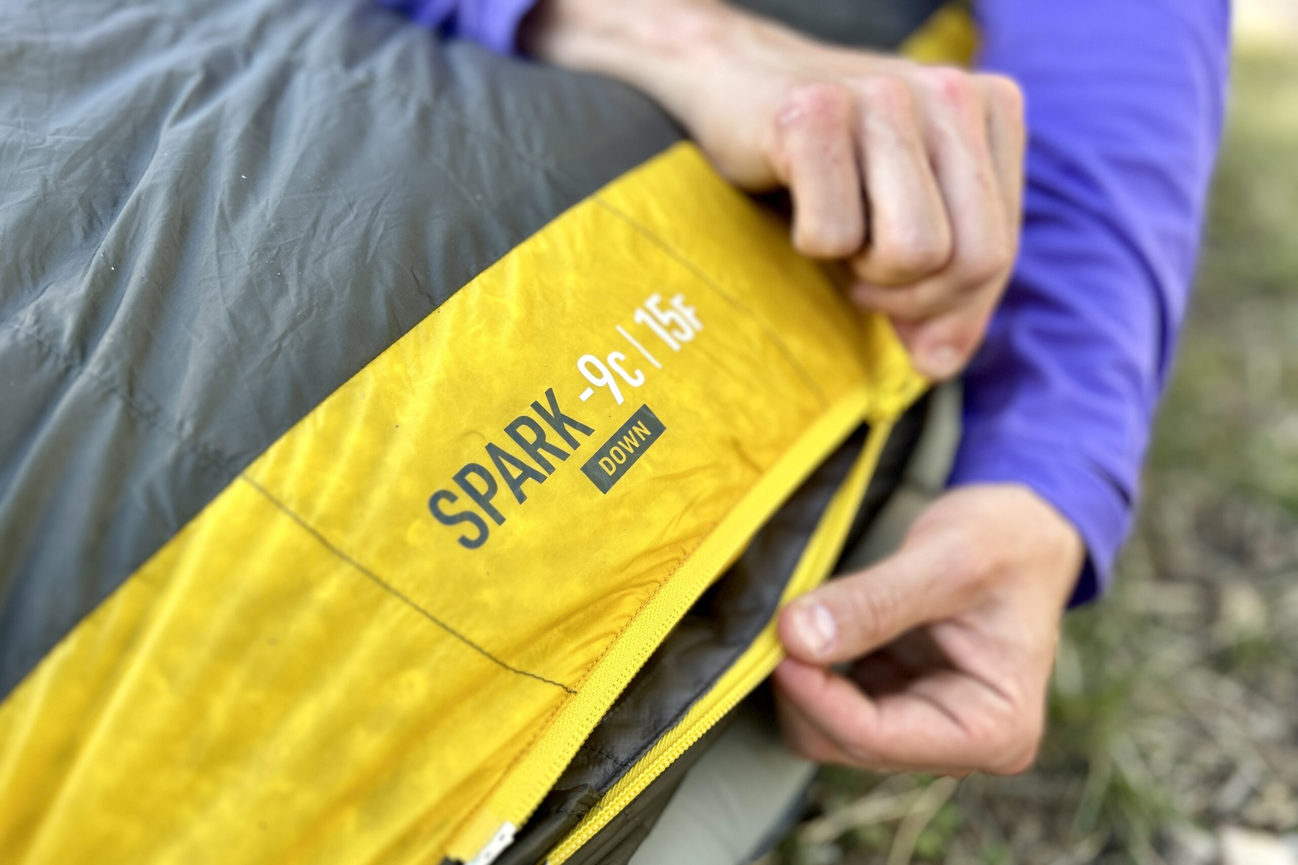 A man zips up a sleeping bag with the logo - Seat to Summit Spark 15 - featured in the center of the image.