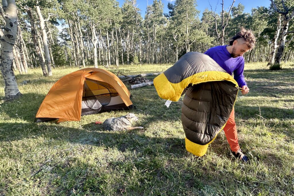 Standing outside of his tent at camp, a man zips up his sleeping bag in the morning light.