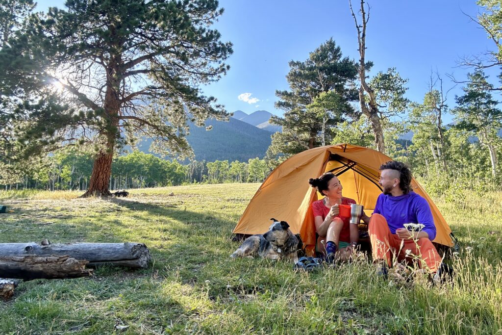 Two people sit outside a yellow tent with forest, blue sky and mountains behind them.