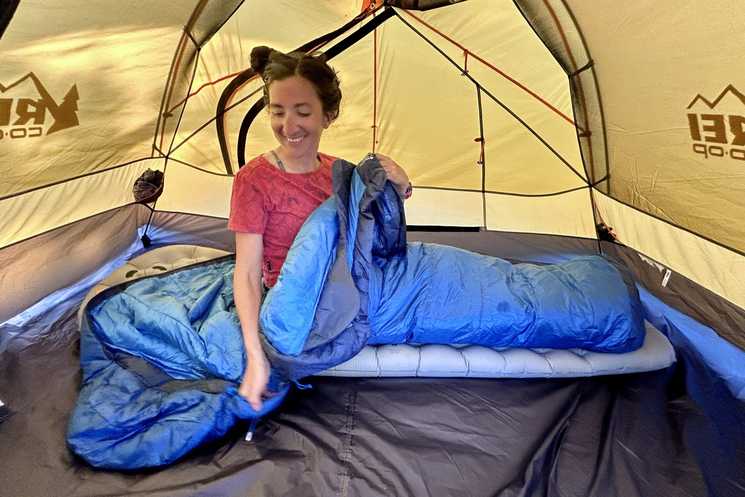 An image of a smiling woman sitting up and adjusting a sleeping bag, on a sleeping pad, in a tent.