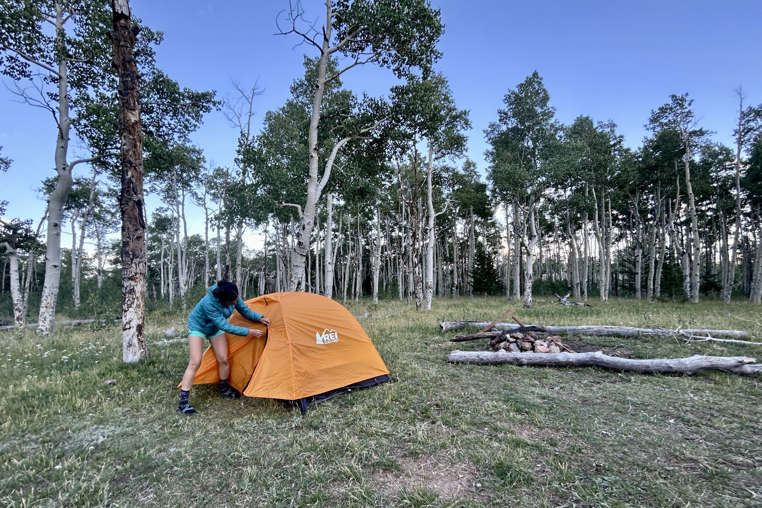 A woman zips up the rainfly of an orange tent in the woods.