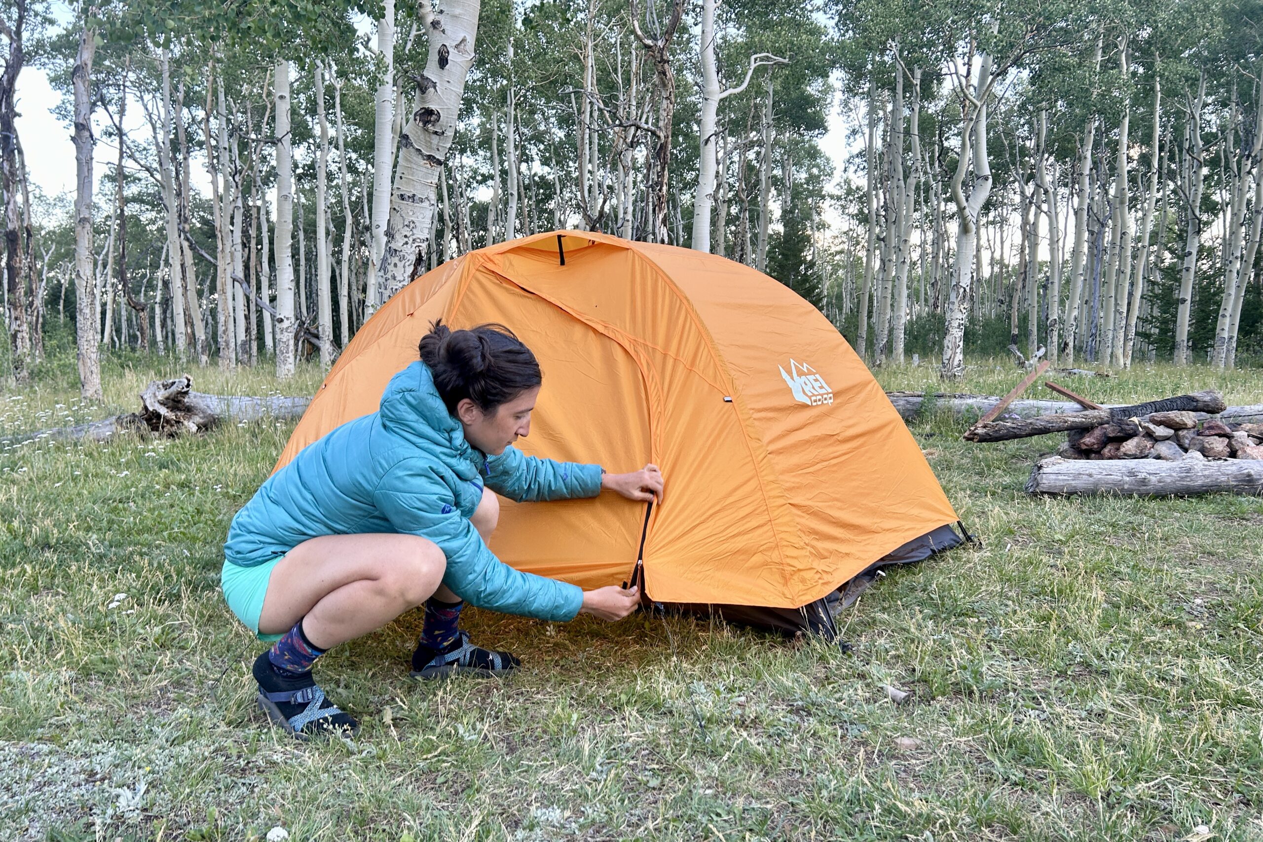 A woman zips up the rainfly of an orange tent in the woods.