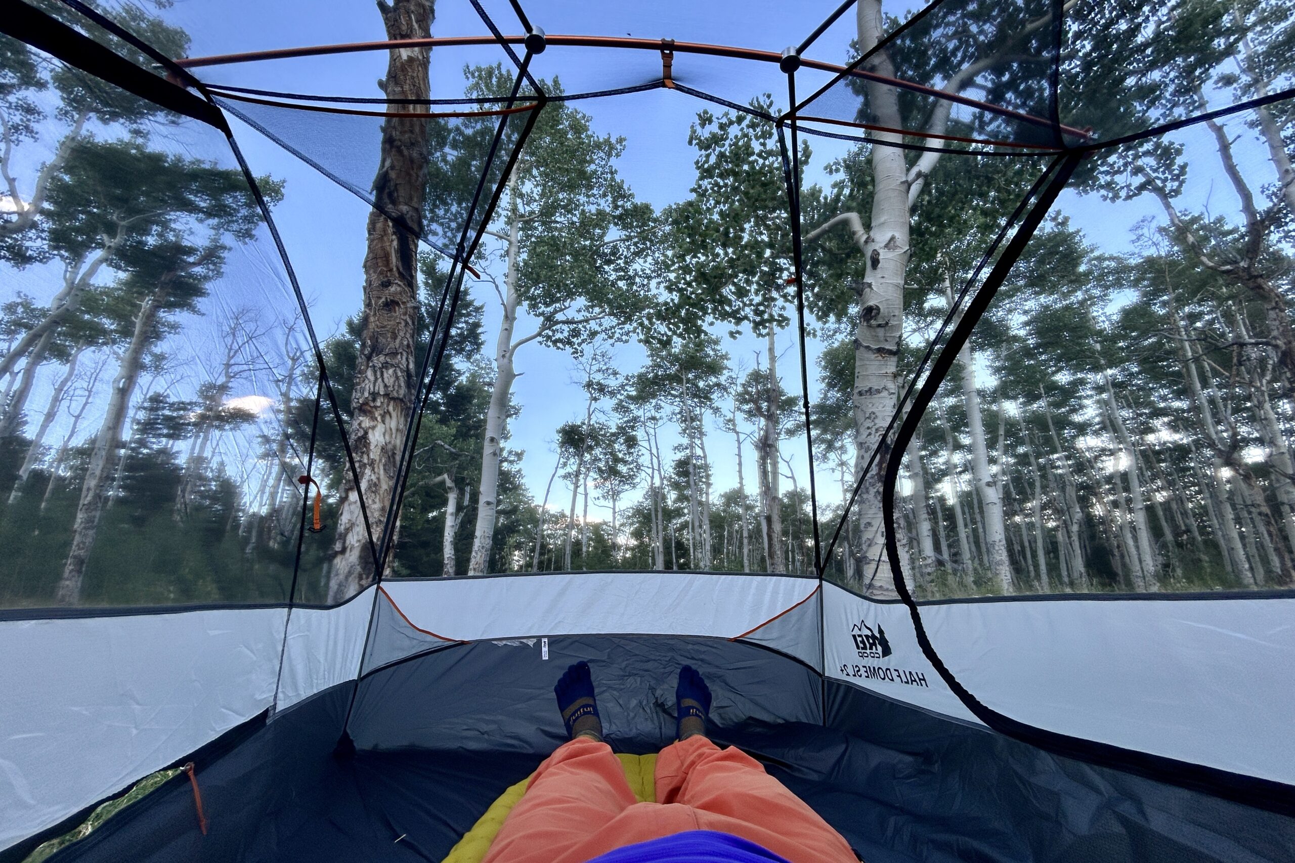 One person from the waist down lays on sleeping pad in a tent with no fly on so you can see the sky and tent poles and mesh.