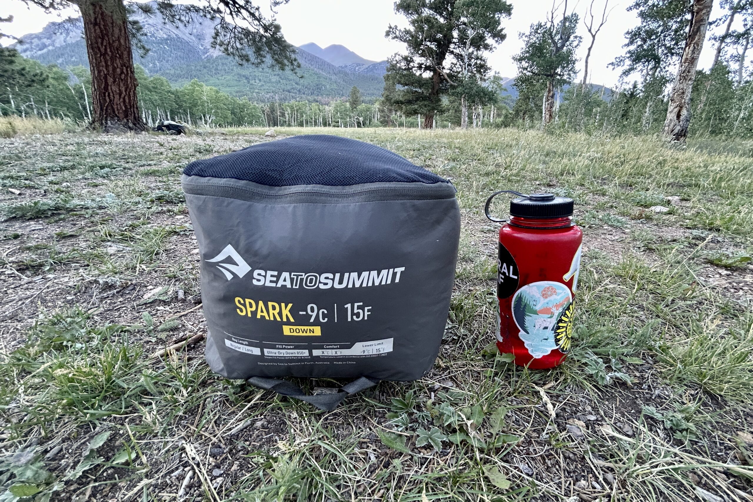 A packed sleeping bag sits next to a water bottle on the ground in a forest.