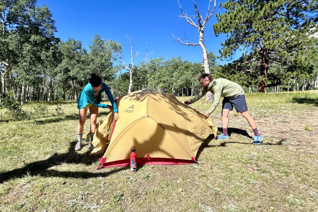 Two people zip up the fly of a tent on a sunny day.