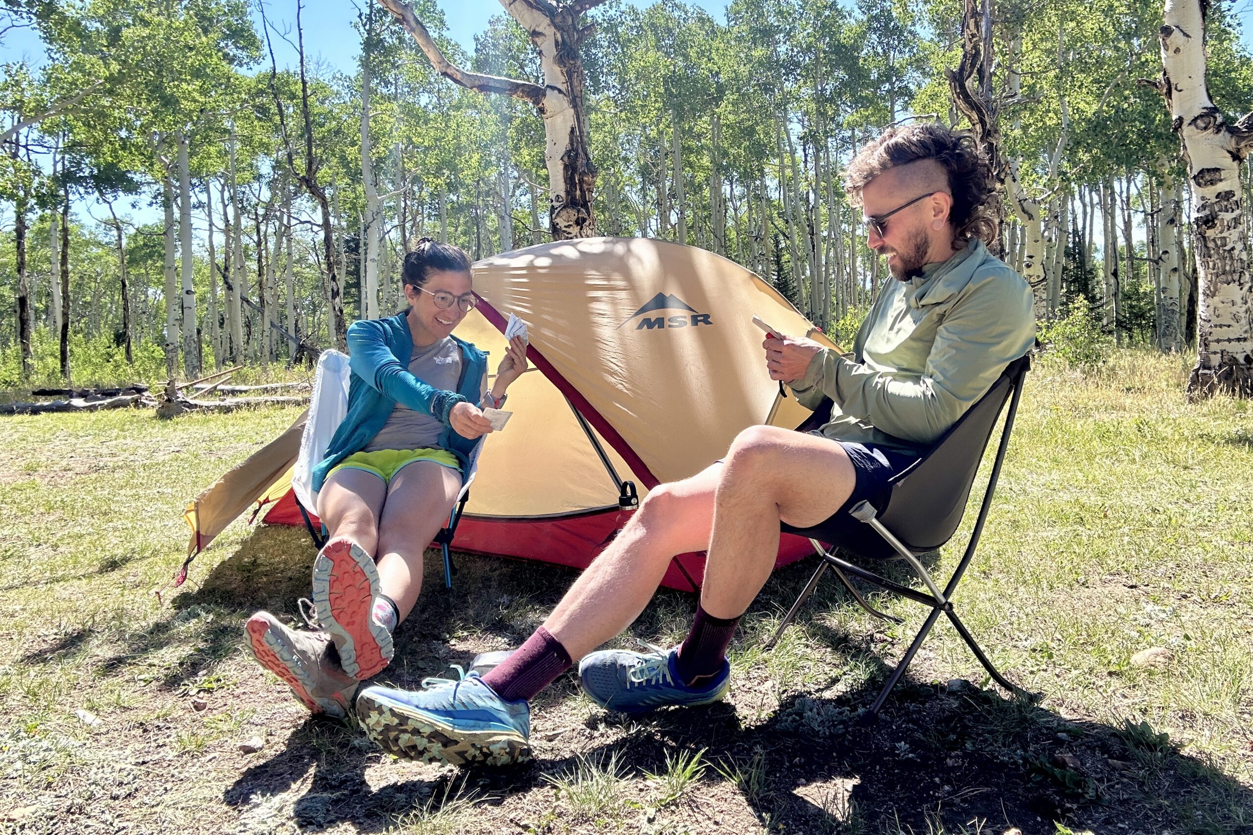 Two people sitting in small camp chairs play cards outside their tent on a sunny day.