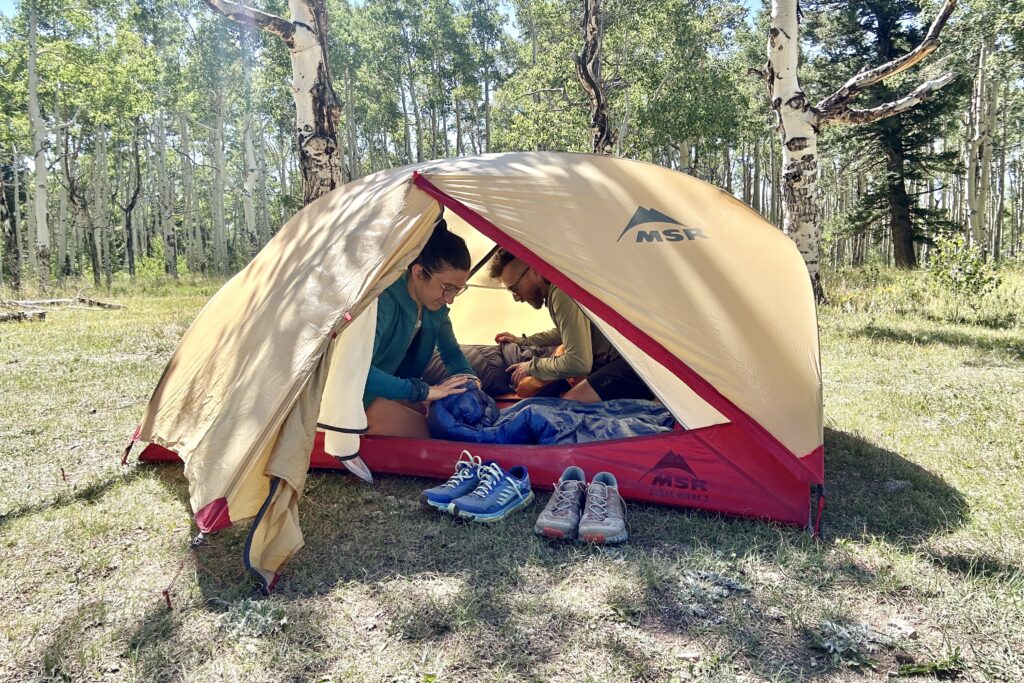 Two people adjust gear inside a tent on a sunny day.