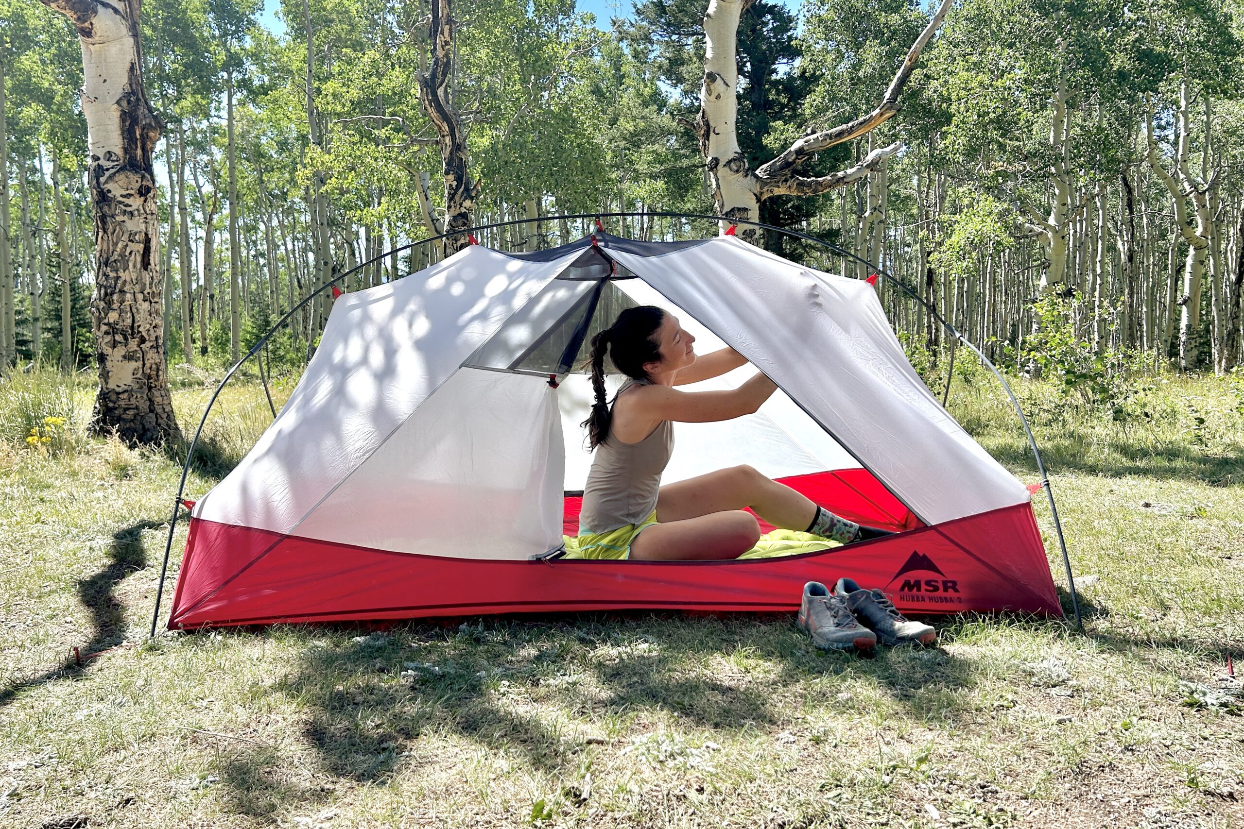 A woman relaxes in a tent with the fly off adjusting the ceiling with forest behind her.