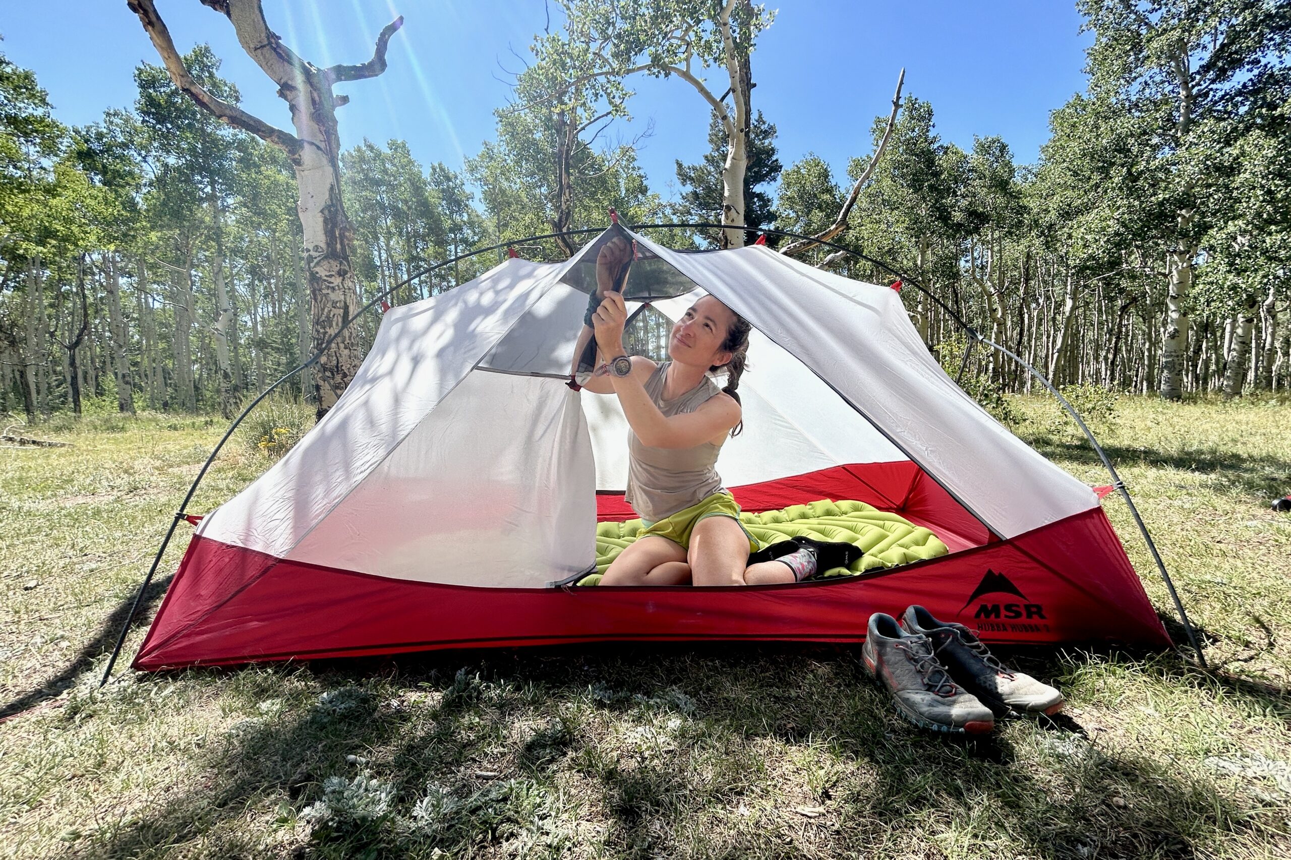 A woman relaxes in a tent adjusting the ceiling with forest behind her.